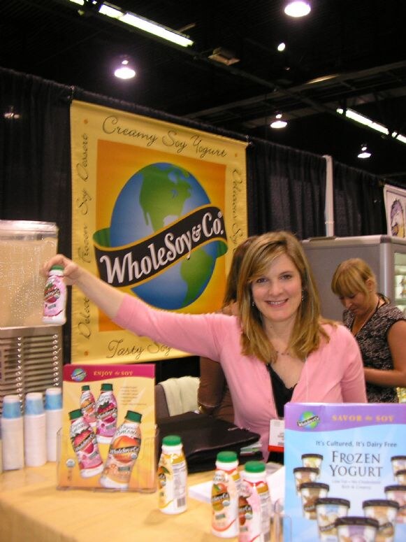 A smiling woman in a pink cardigan holds up a bottle of WholeSoy & Co. soy yogurt drink beside a yellow banner. Bottles and a Frozen Yogurt display sit on the table.