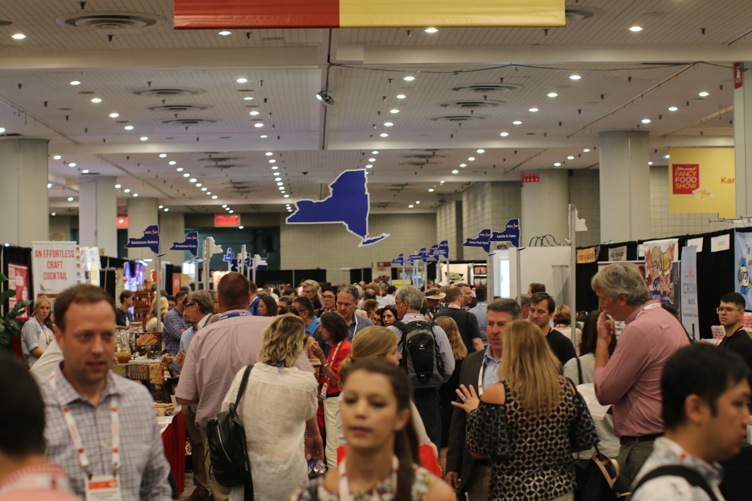 Dense crowd of badge-wearing attendees in an exhibit aisle with blue New York-shaped signs and booths on both sides.