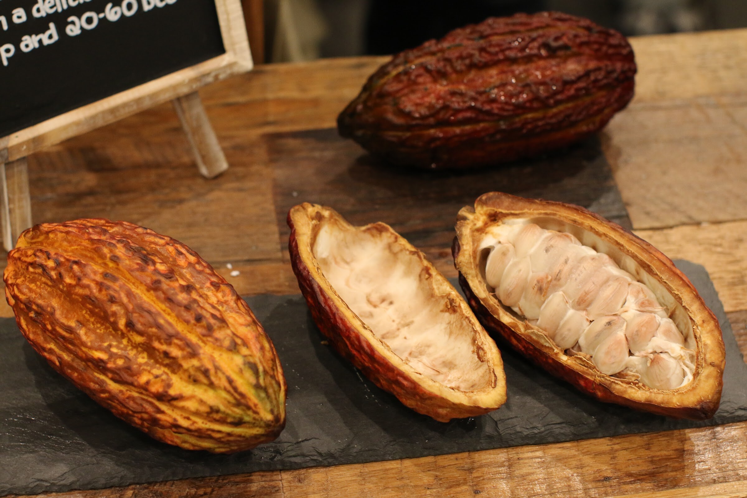 Three cacao pods, one split open to show white pulp and beans, on a slate slab on a wooden table.