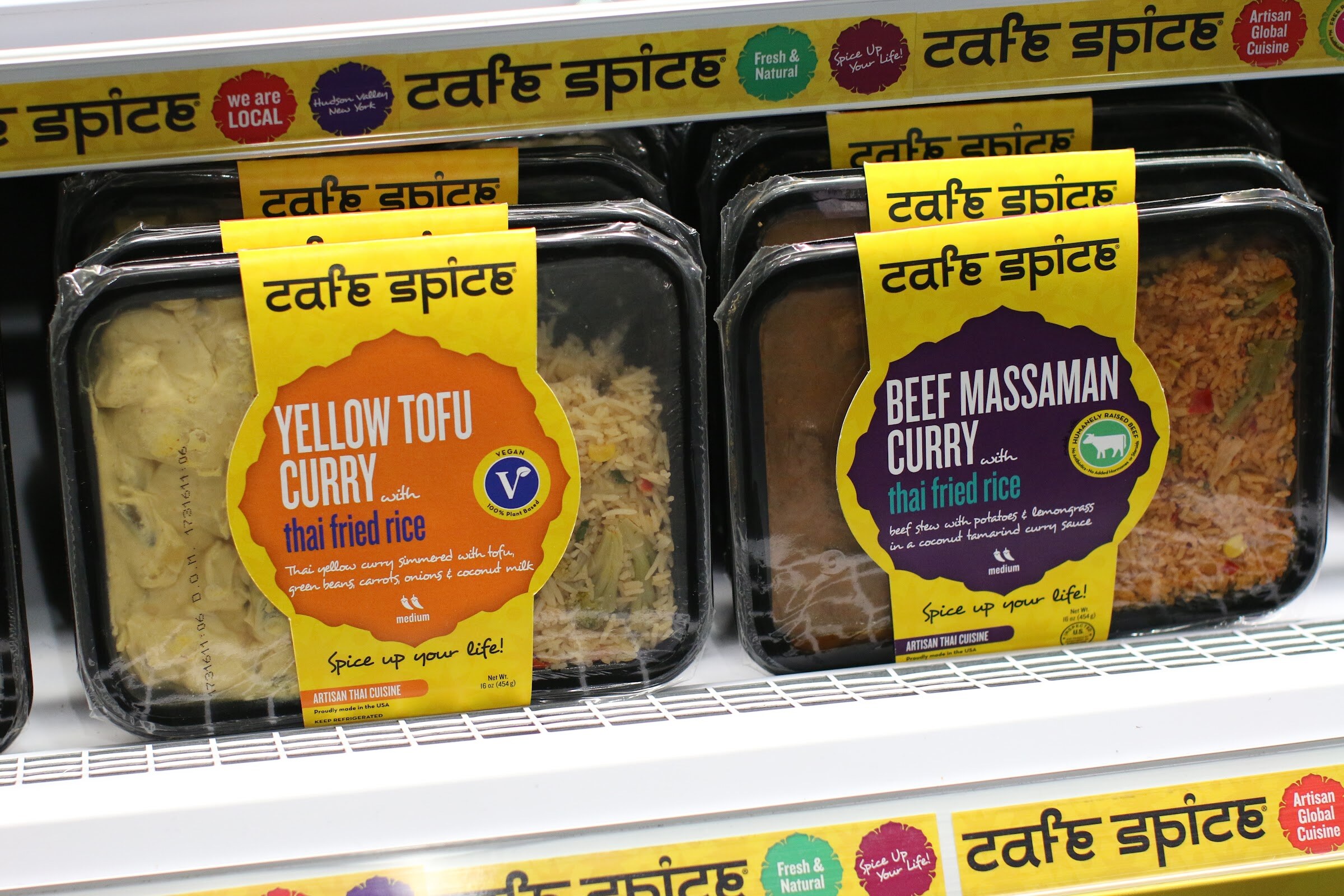 Two black trays of Cafe Spice prepared meals with yellow and purple labels on a cooler shelf.