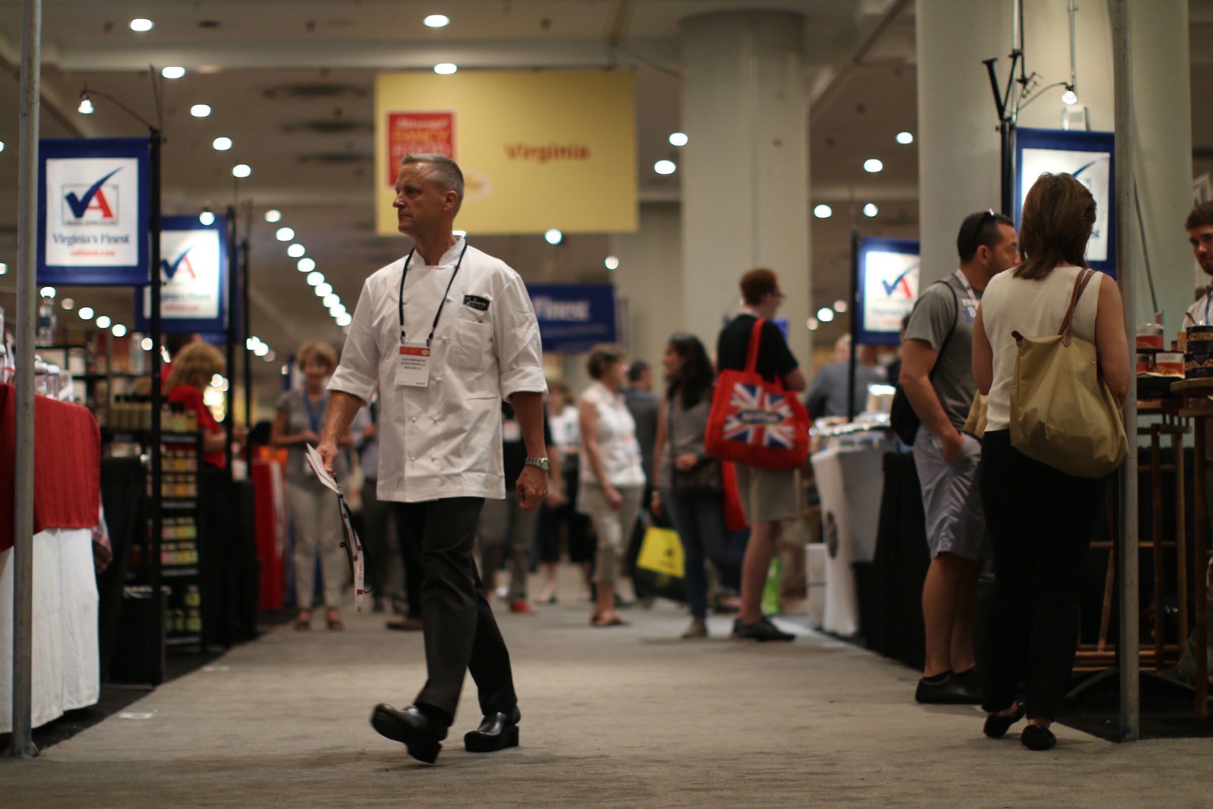 Man in a white chef coat and lanyard walking along a carpeted aisle with Virginia's Finest signs and other attendees behind him.