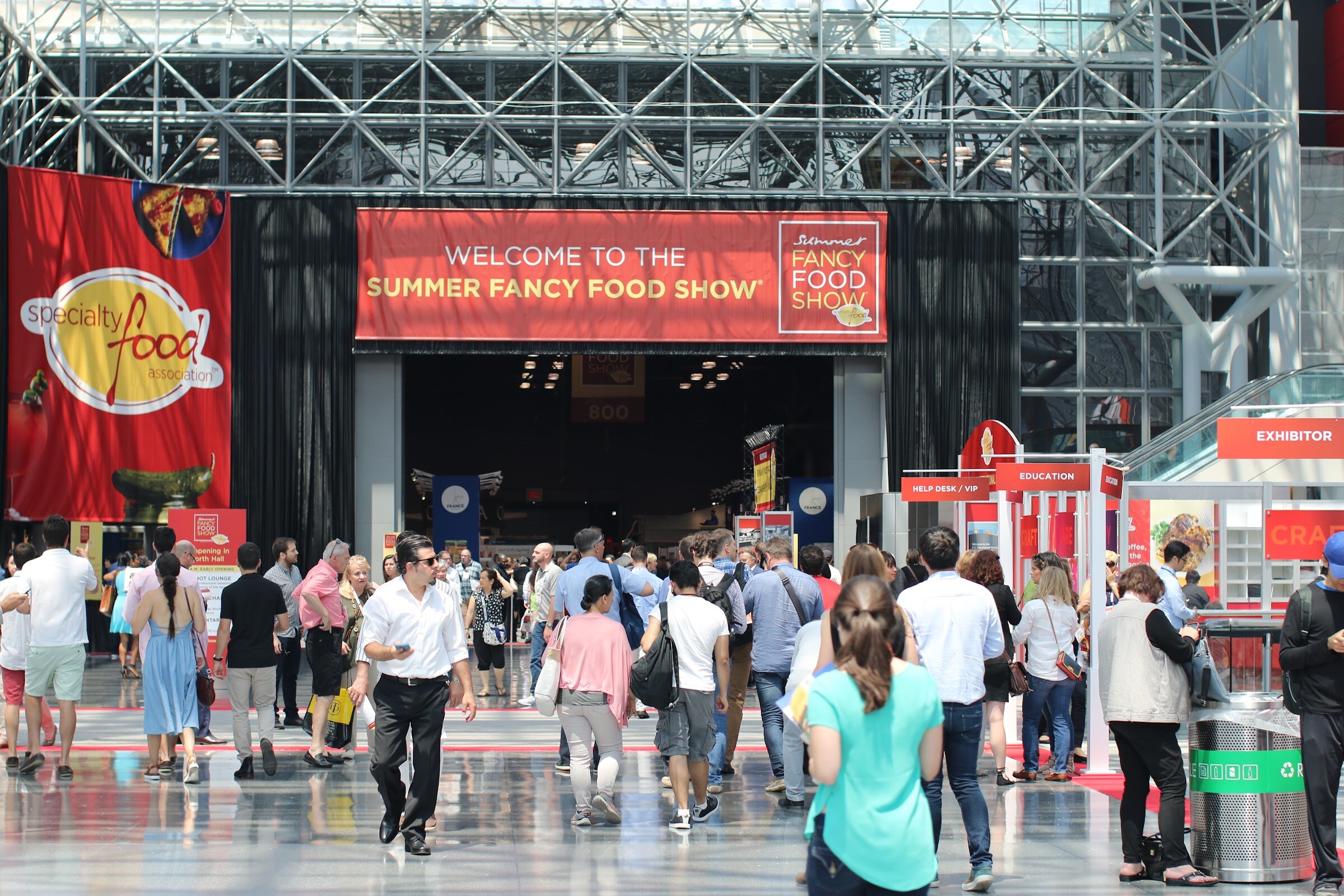 Busy crowd of people walking toward an entrance with a red welcome banner and a Specialty Food Association banner.