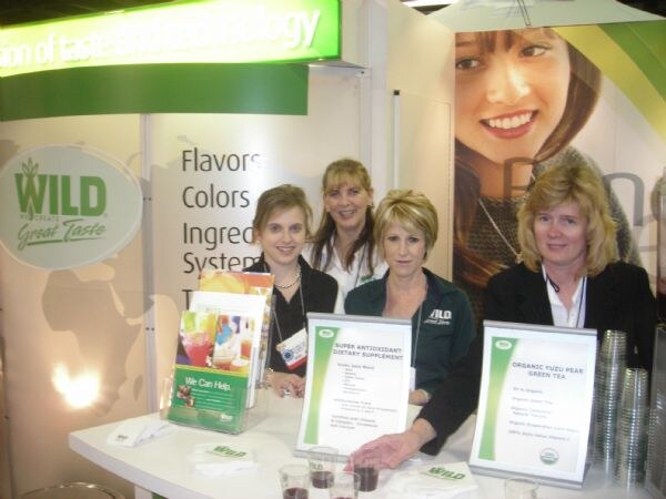 Four women stand behind a curved counter at the Wild Flavors booth, with brochures, two signs on antioxidant supplement and organic yuzu pear green tea, and cups of drink.
