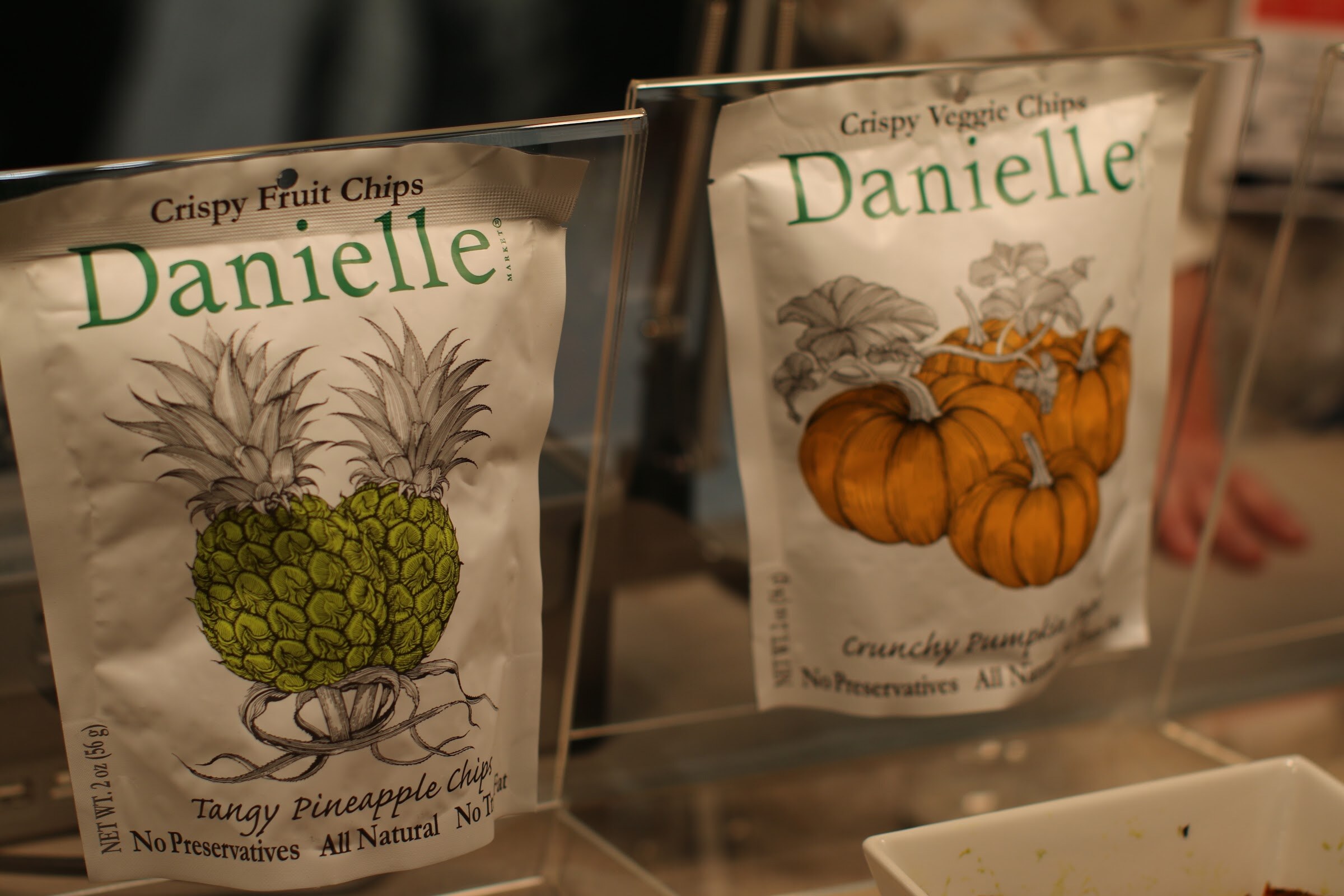 Two white Danielle chip pouches, pineapple and pumpkin, displayed on clear acrylic stands.
