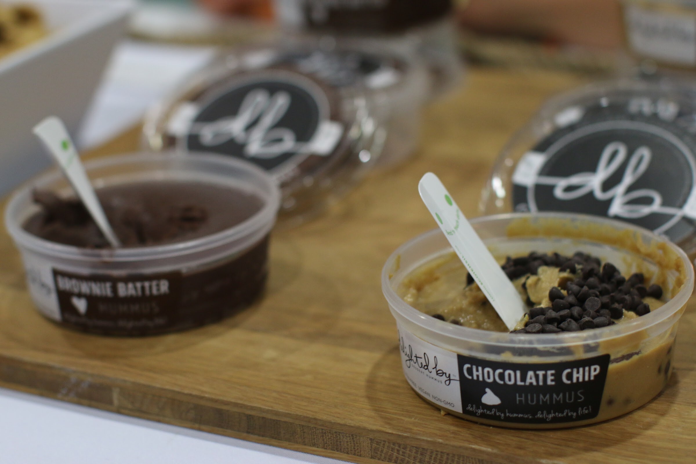 Two tubs of Delighted By hummus, brownie batter and chocolate chip, with tasting spoons on a wooden board.