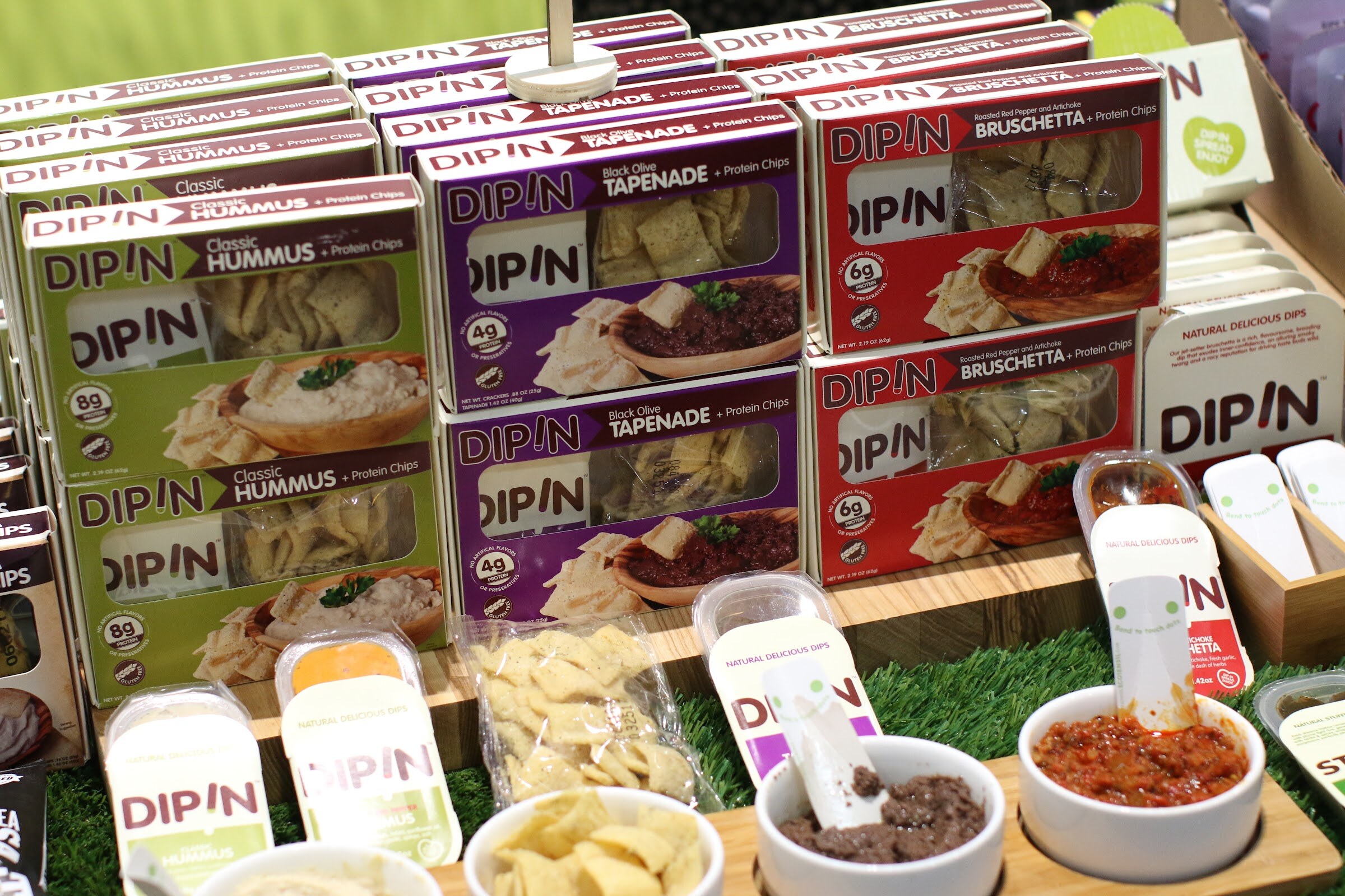 Display of DIP!N boxes in green, purple and red above small bowls of hummus, tapenade and tomato dip.