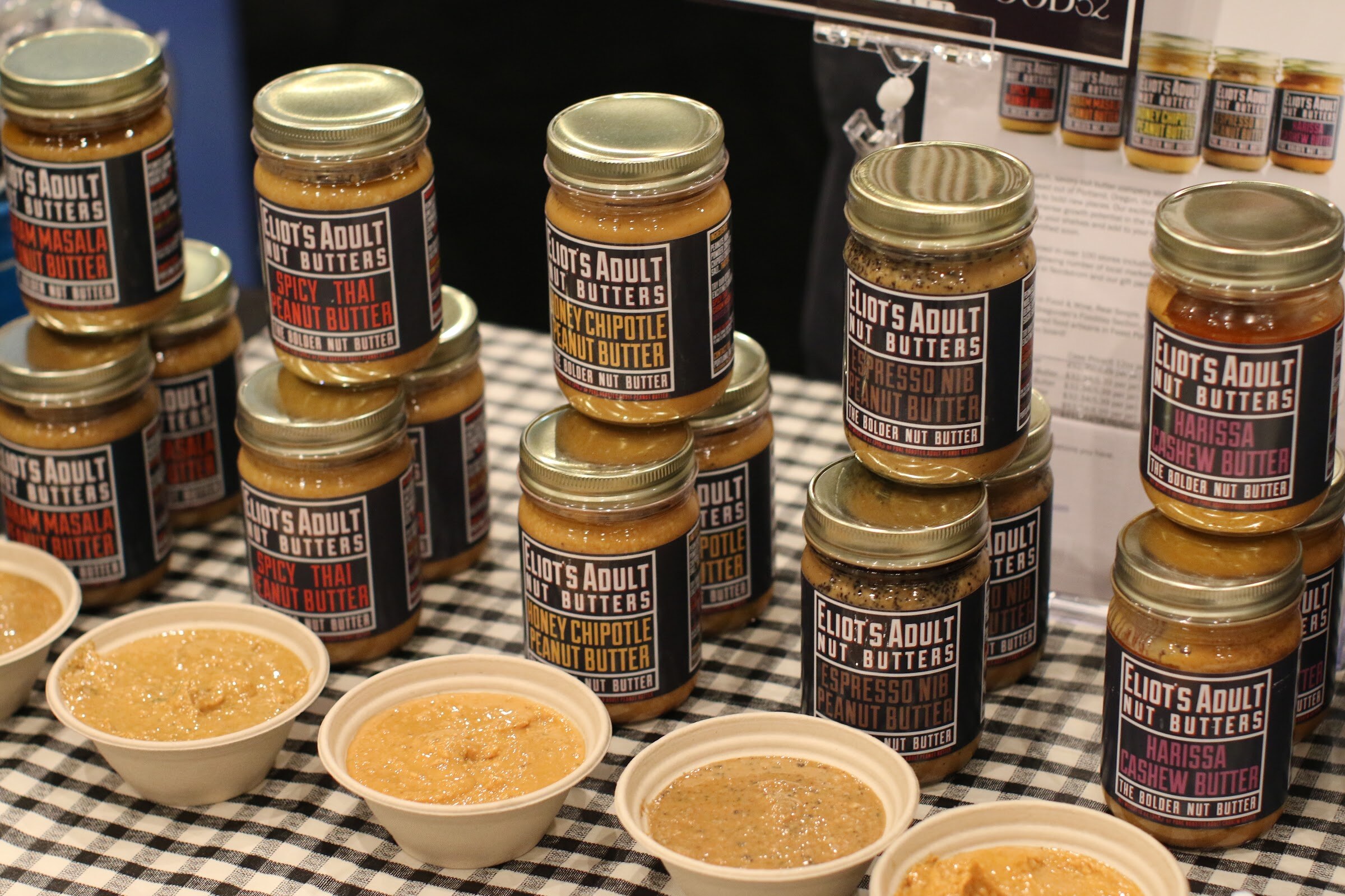 Stacks of Eliot's Adult Nut Butters jars in several flavors above small bowls of sample nut butter.