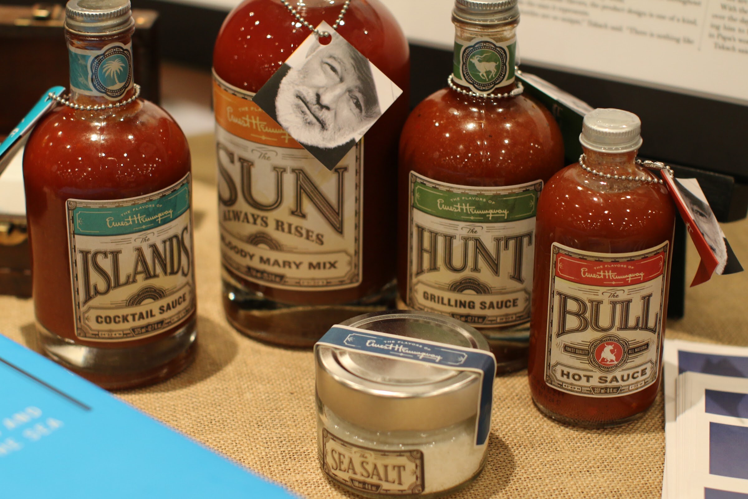 Four bottles of red sauces with vintage labels and portrait tags, beside a small jar of sea salt on burlap.