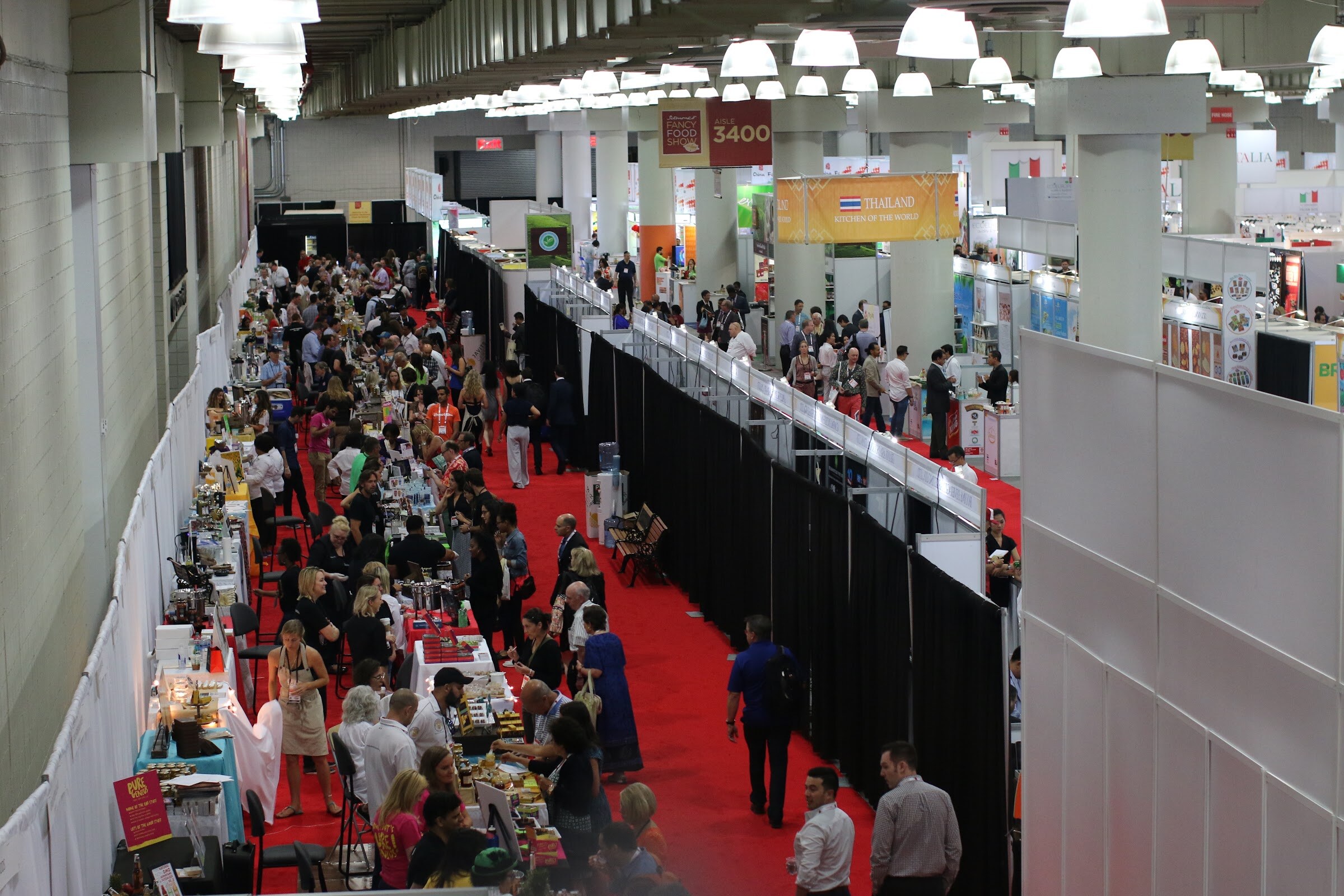 Aerial view of a long aisle with red carpet, crowded booths on the left and black drapes on the right.