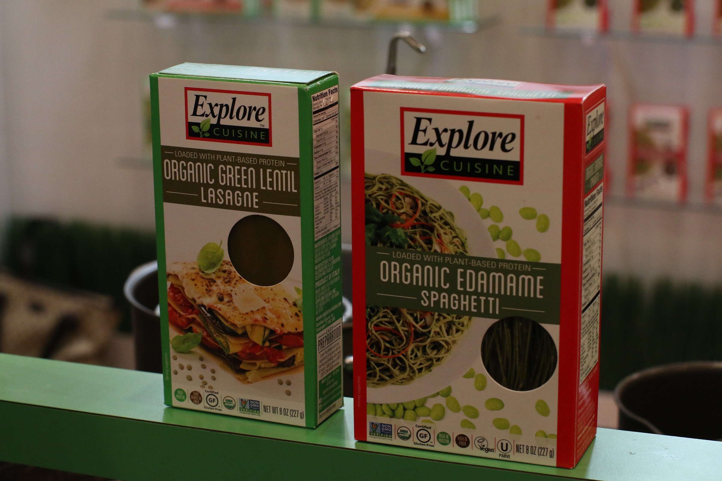 Two boxes of Explore Cuisine pasta, green lentil lasagne and edamame spaghetti, on a green shelf.