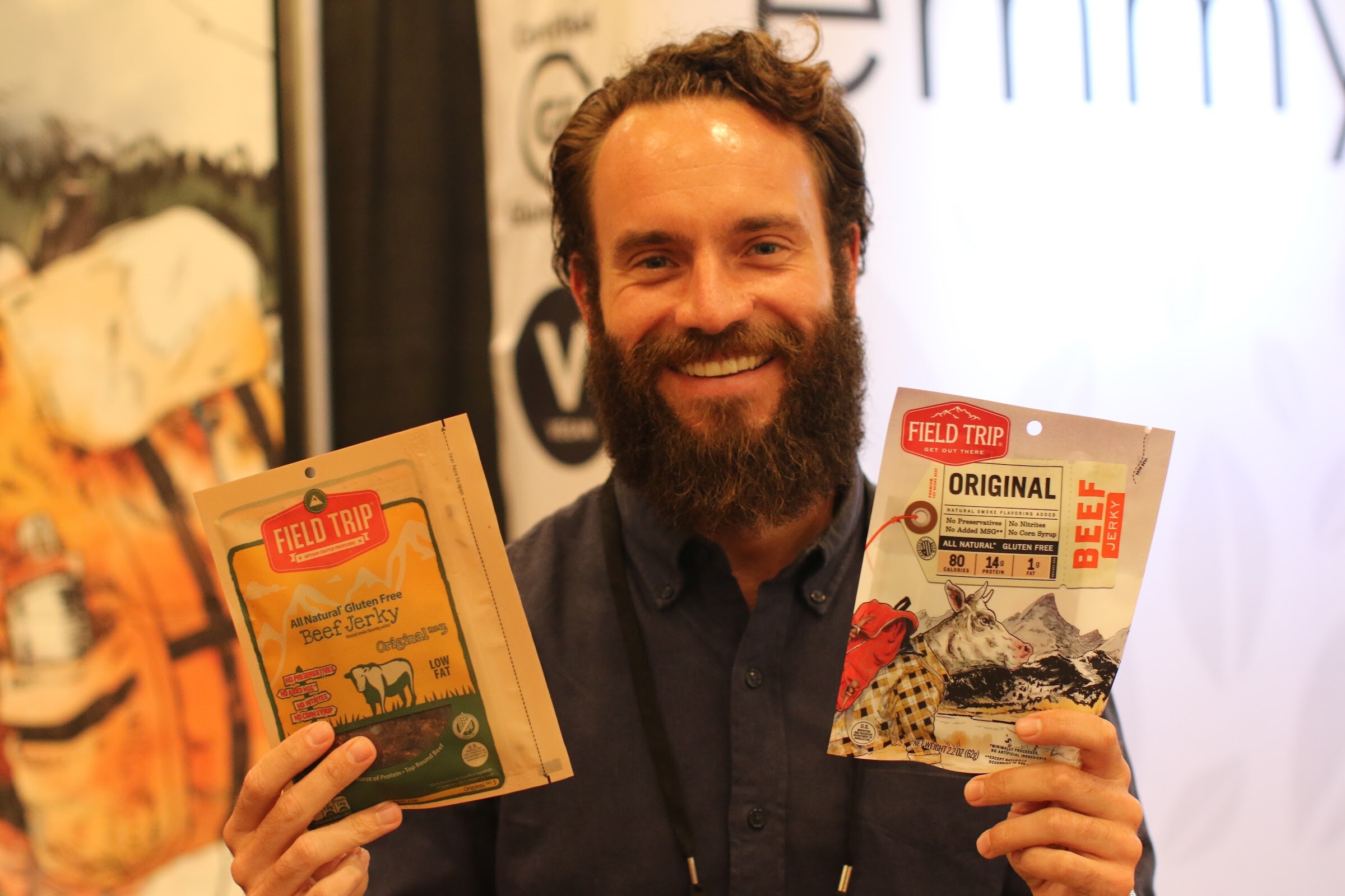 Bearded man smiling and holding a bag of Field Trip beef jerky in each hand in front of a booth backdrop.