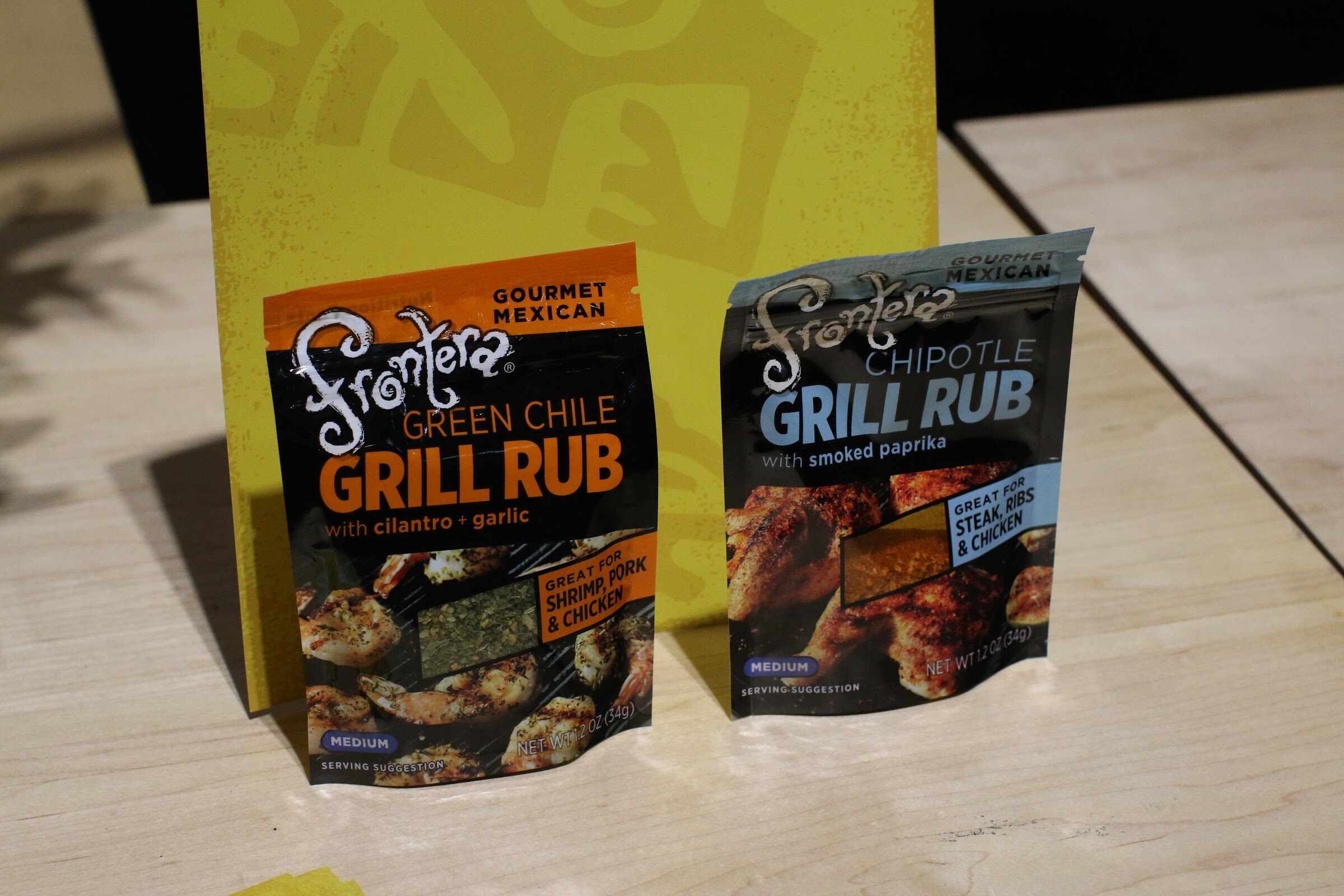 Two Frontera grill rub pouches on a light wood table in front of a yellow sign.