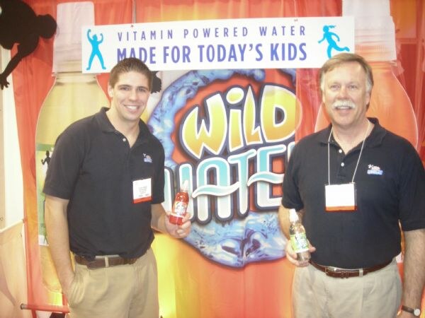 Two men in dark polo shirts and badges hold small bottles in front of a Wild Water backdrop with a sign reading Vitamin Powered Water, Made for Today's Kids.