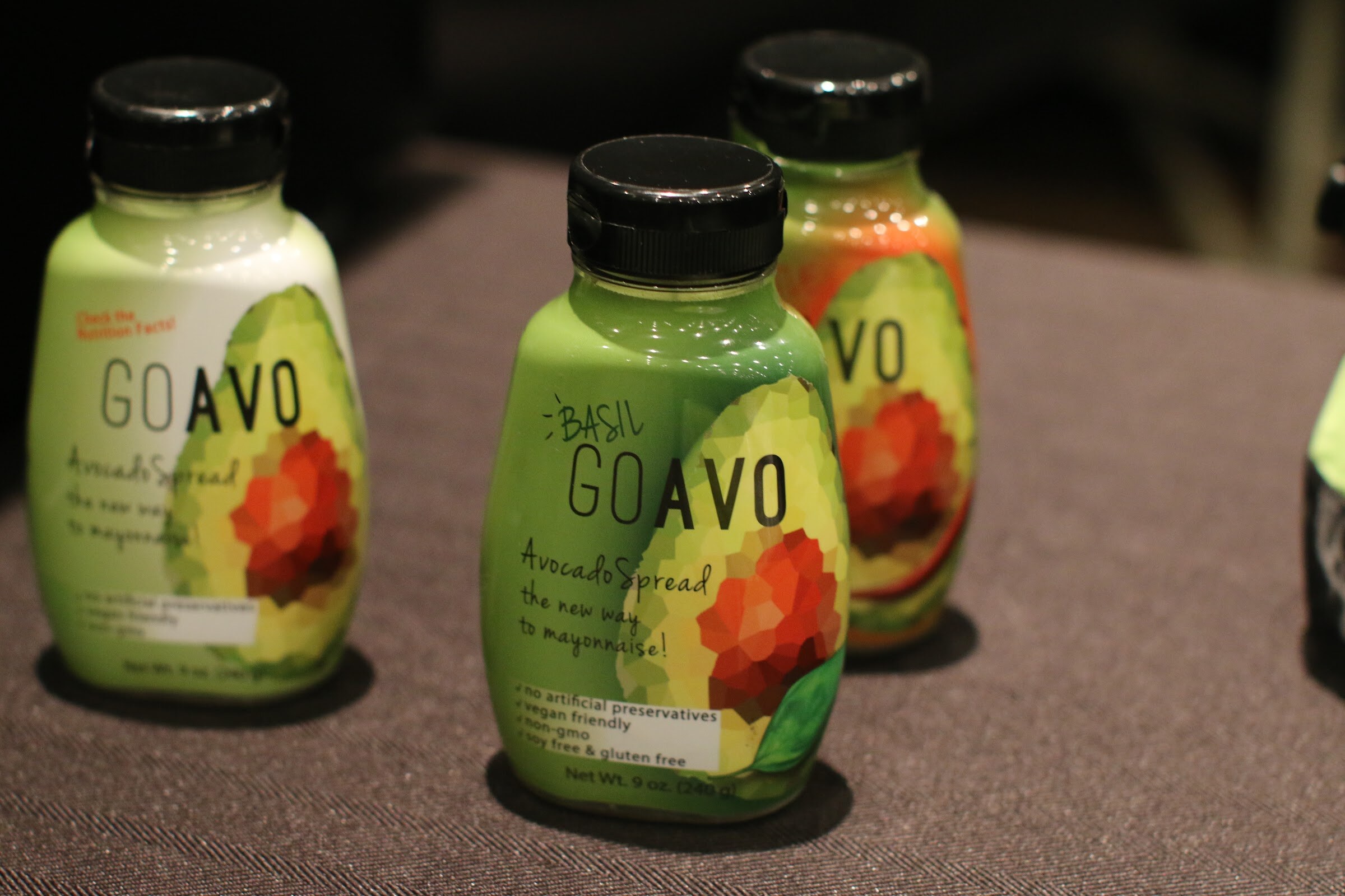 Three green GoAvo avocado spread bottles with black caps on a brown table.