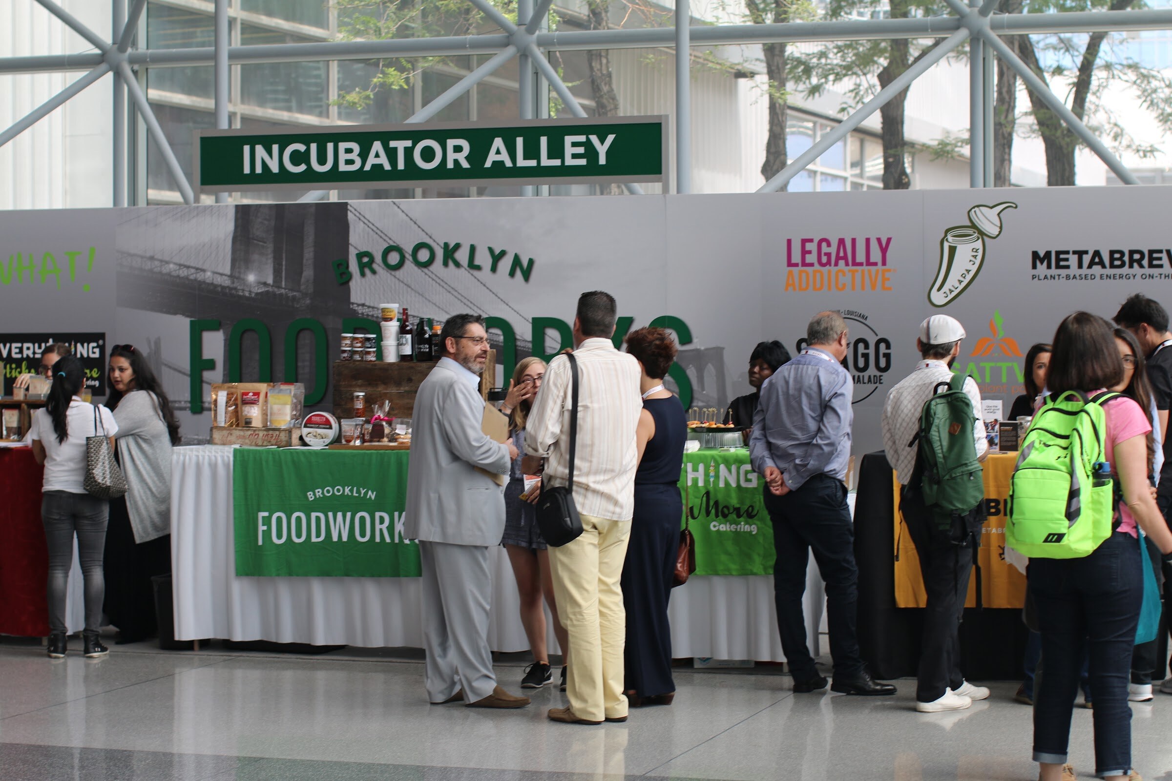 People standing at tables under an Incubator Alley sign, with Brooklyn Foodworks banners and glass roof framing behind them.