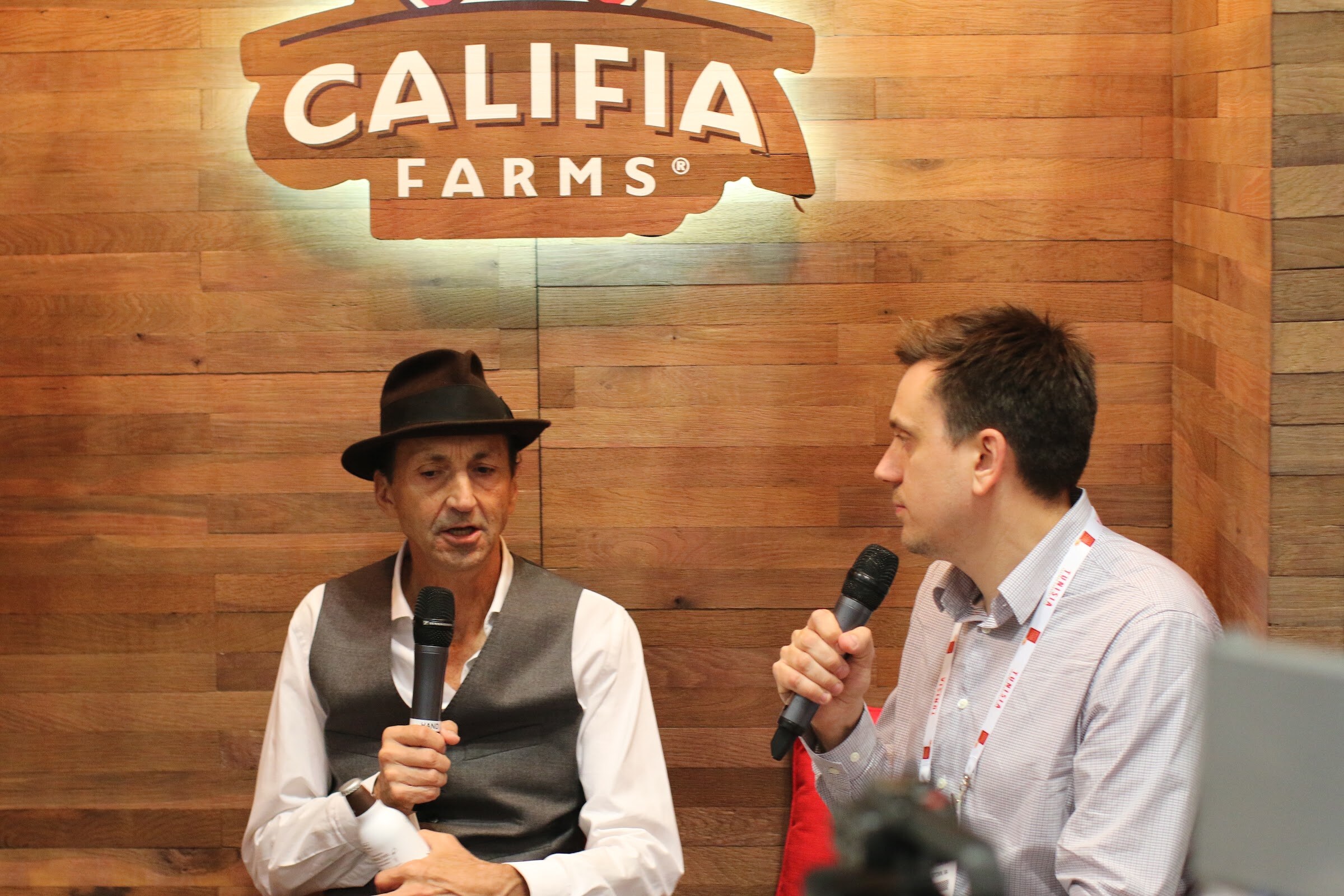 Two seated men with microphones talking in front of a wooden wall with a glowing Califa Farms logo.