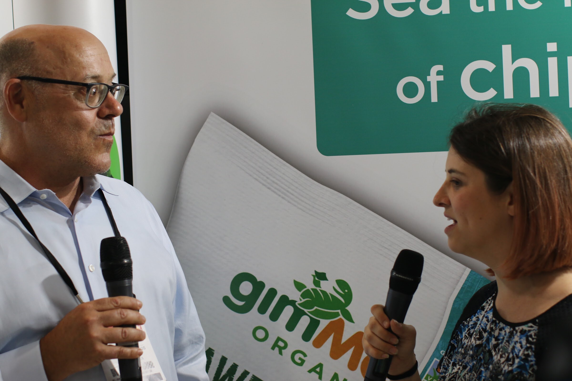 Man with glasses and a woman each holding microphones, facing each other in front of a gimMe Organic banner.