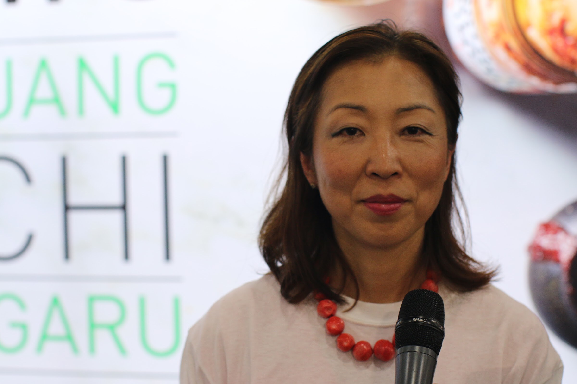 Woman with shoulder-length brown hair and a red bead necklace holding a microphone, with partial large lettering behind her.