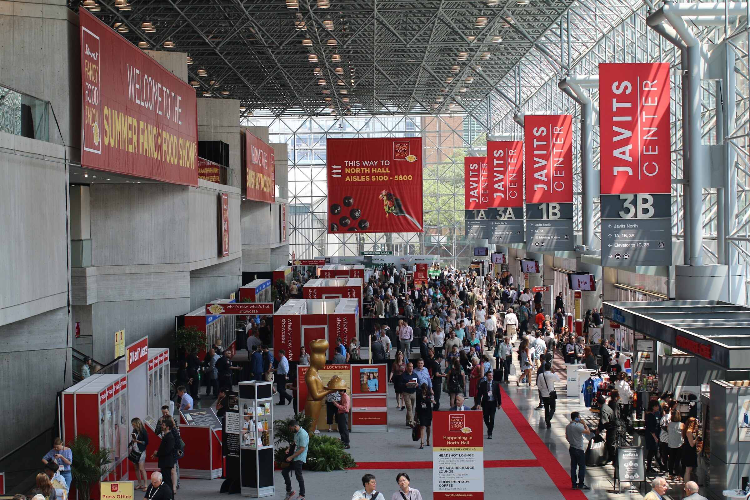 Elevated view of a long glass-roofed convention concourse filled with people, with red Javits Center banners hanging overhead.