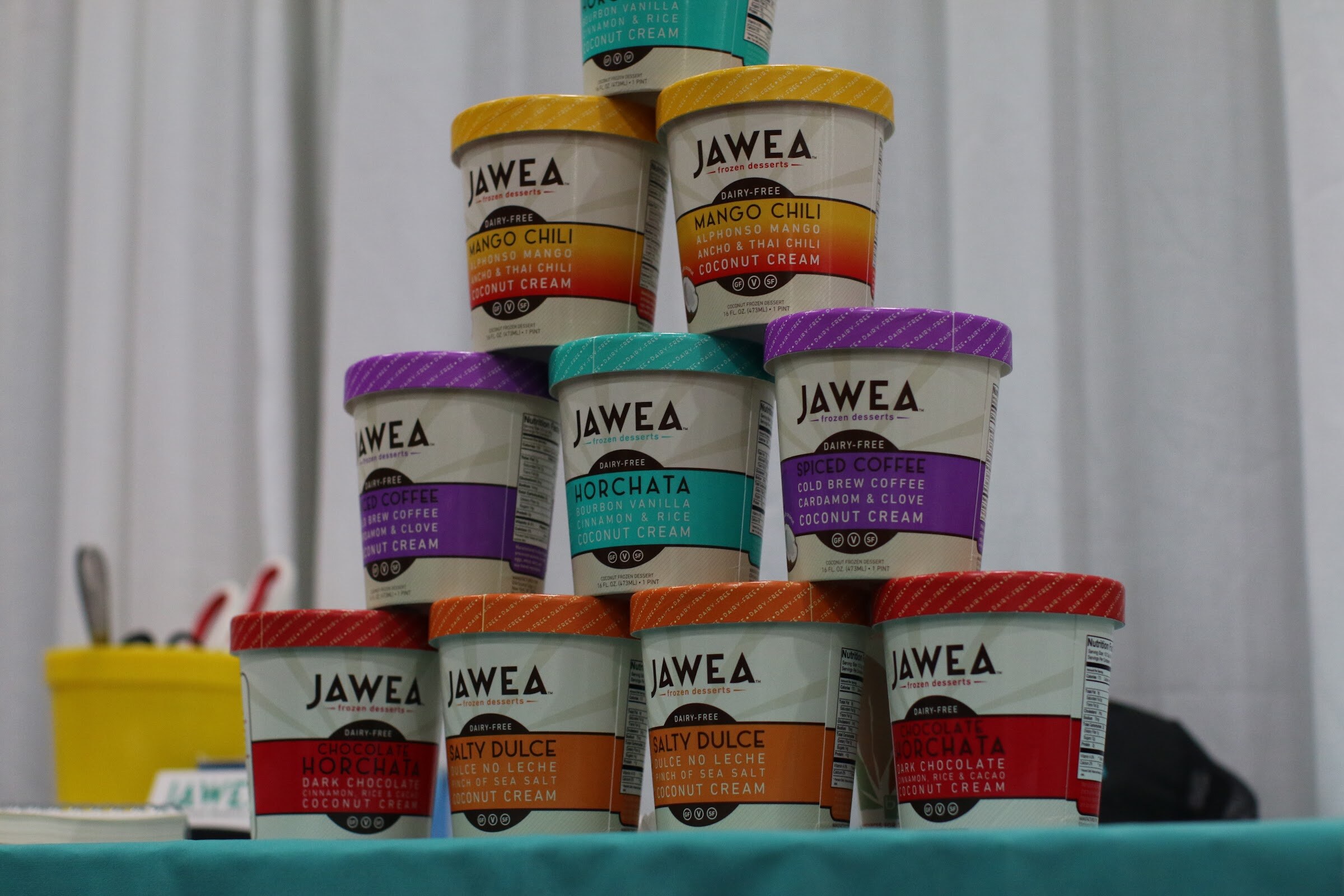 Pyramid of JAWEA frozen dessert pints with colored lids stacked on a teal tablecloth in front of a white curtain.