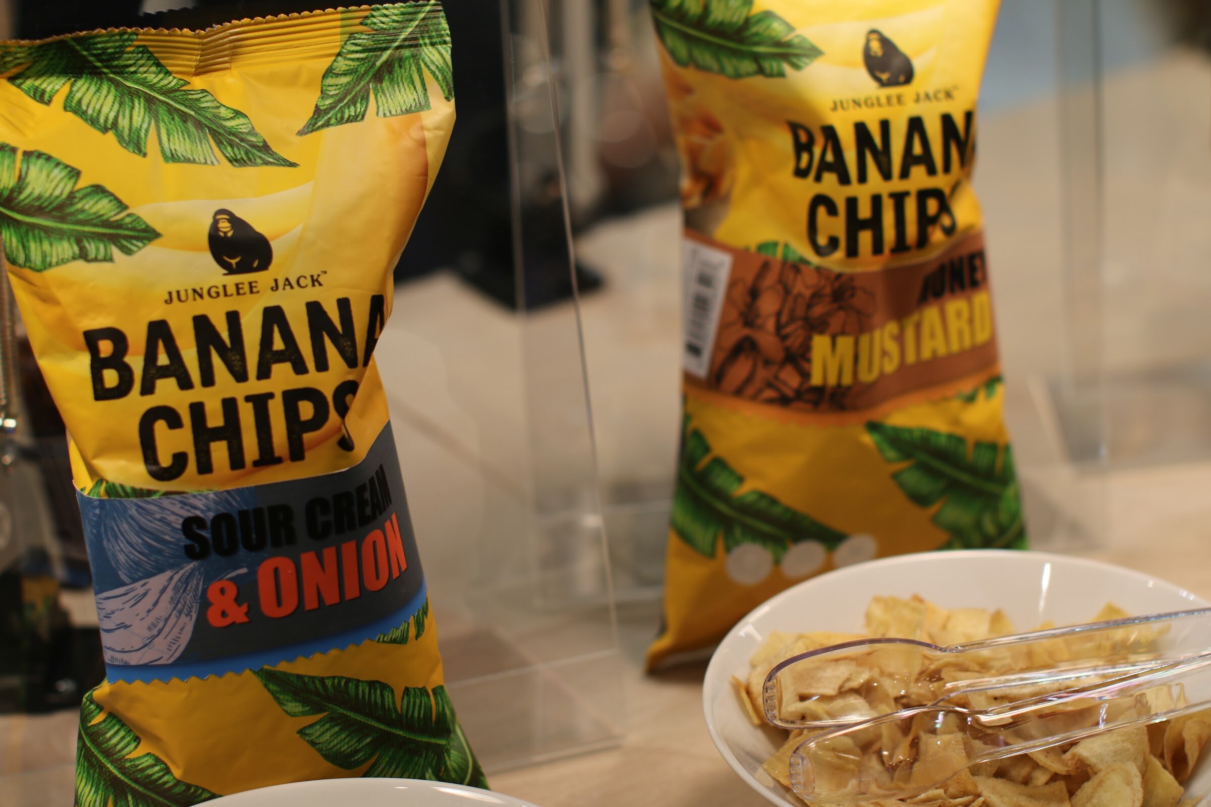 Two bags of Junglee Jack banana chips next to a white bowl of chips with clear tongs.