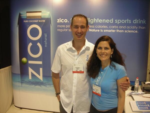 A man in a white shirt with his arm around a woman in a light blue Zico shirt, both wearing badges, in front of a Zico pure coconut water display.