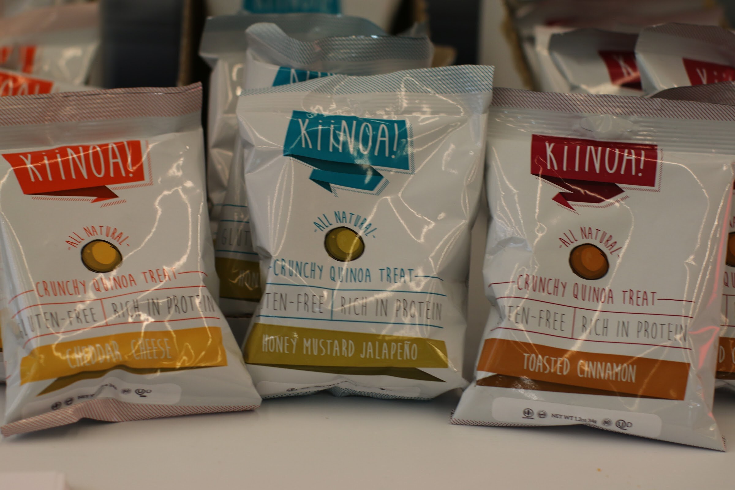 Three bags of Kiinoa all natural crunchy quinoa treats in white packaging with colored banners.