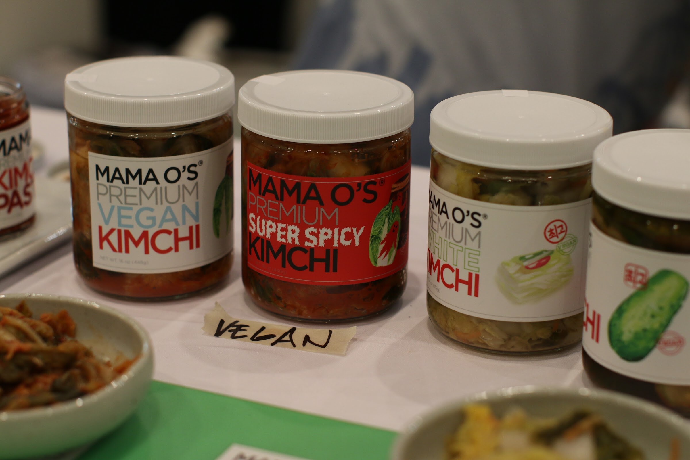 Four jars of Mama O's kimchi with white lids and a taped VEGAN label on a white table.
