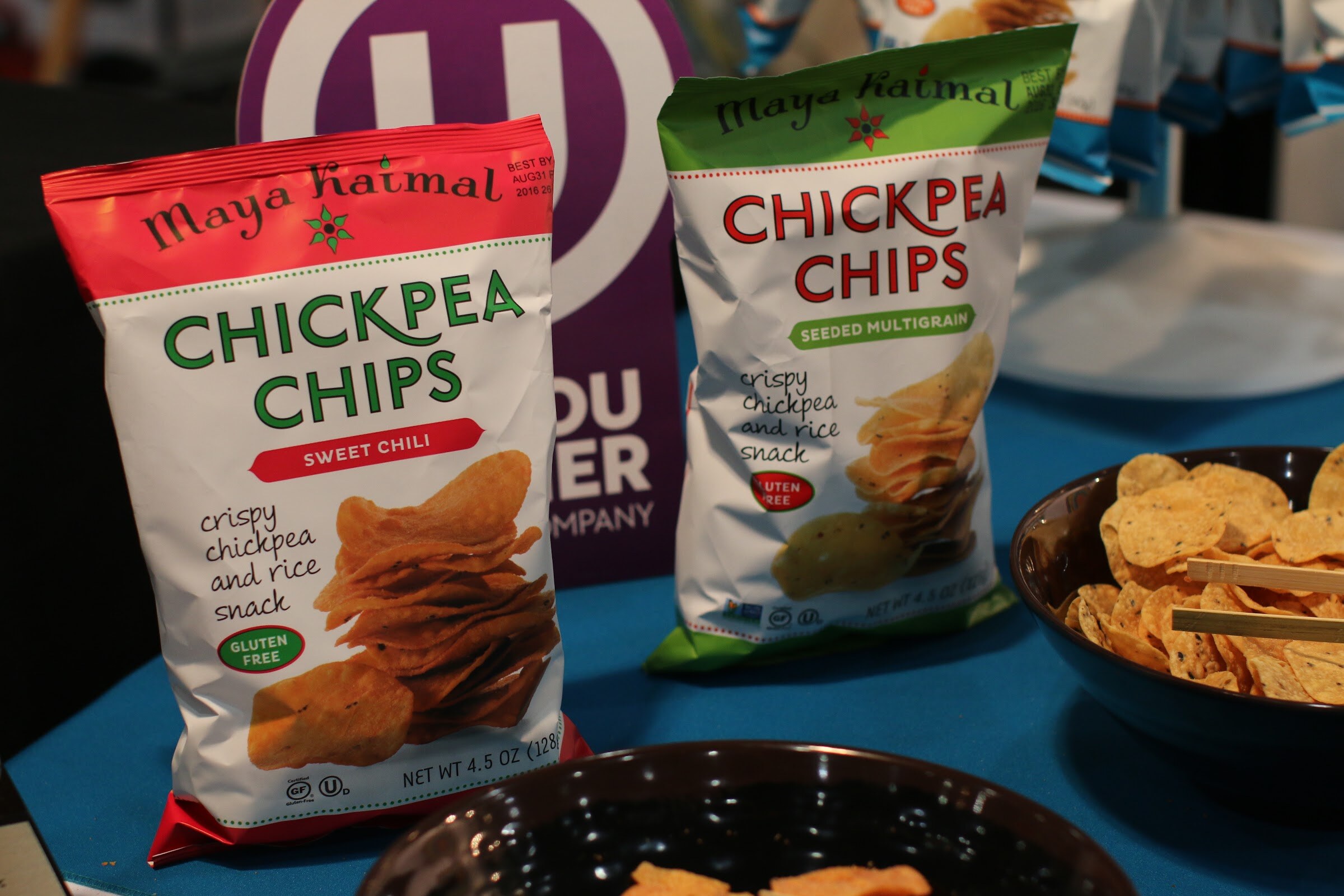 Two bags of Maya Kaitmal chickpea chips on a teal tablecloth with bowls of tortilla-style chips.