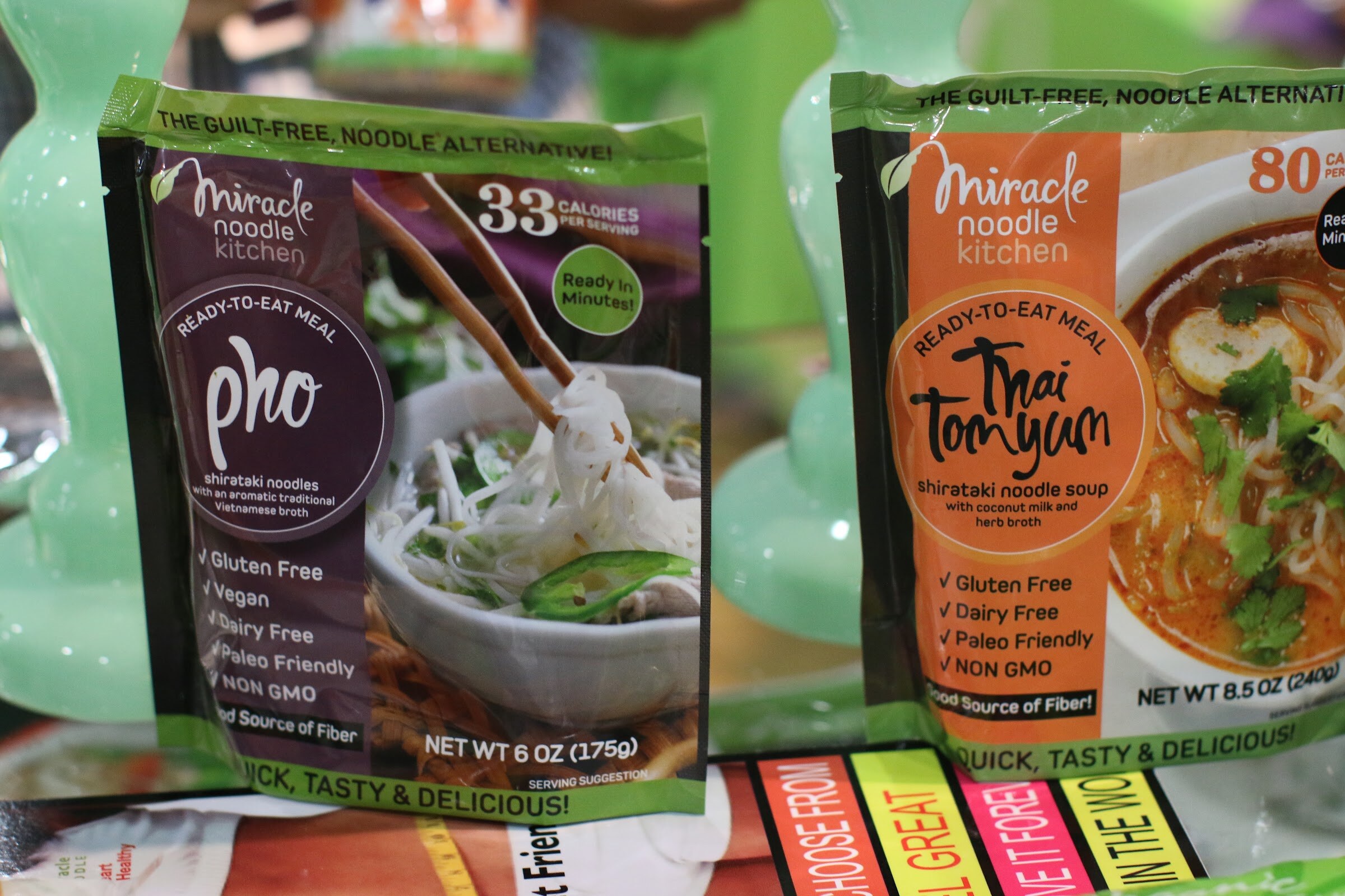 Two Miracle Noodle Kitchen pouches, purple pho and orange Thai tom yum, in front of green candlestick-style props.