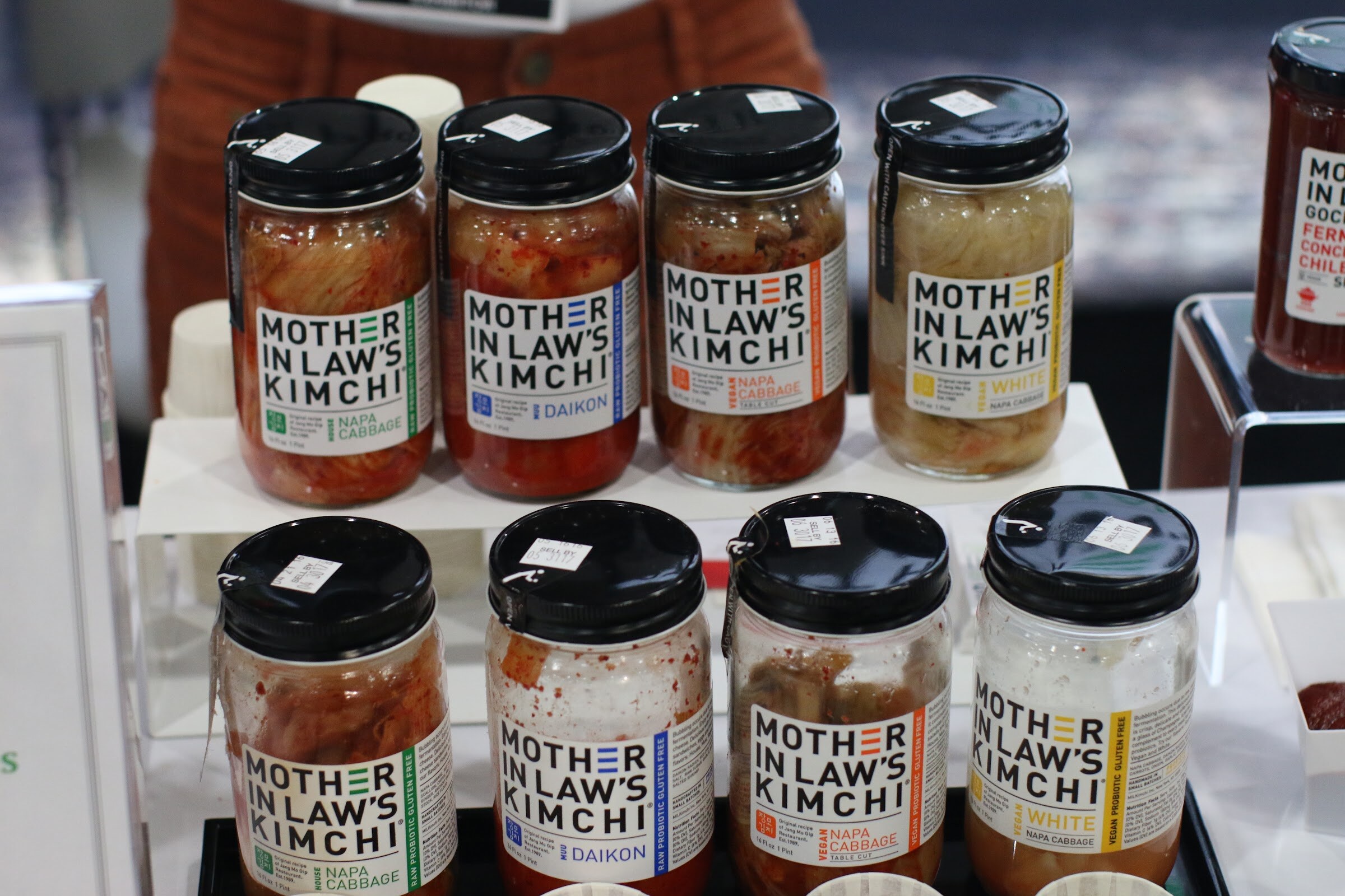 Two rows of Mother in Law's Kimchi jars with black lids on a white riser.