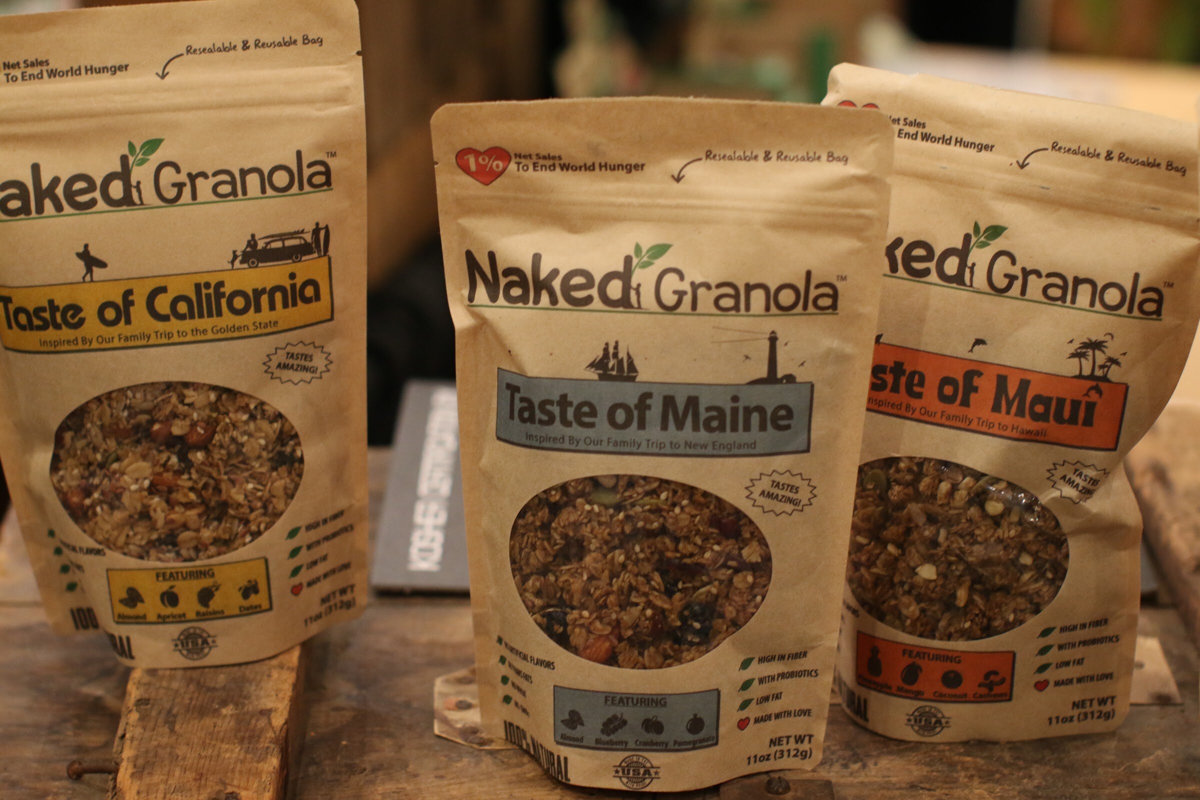Three kraft paper Naked Granola bags with clear windows showing granola, on a weathered wooden table.