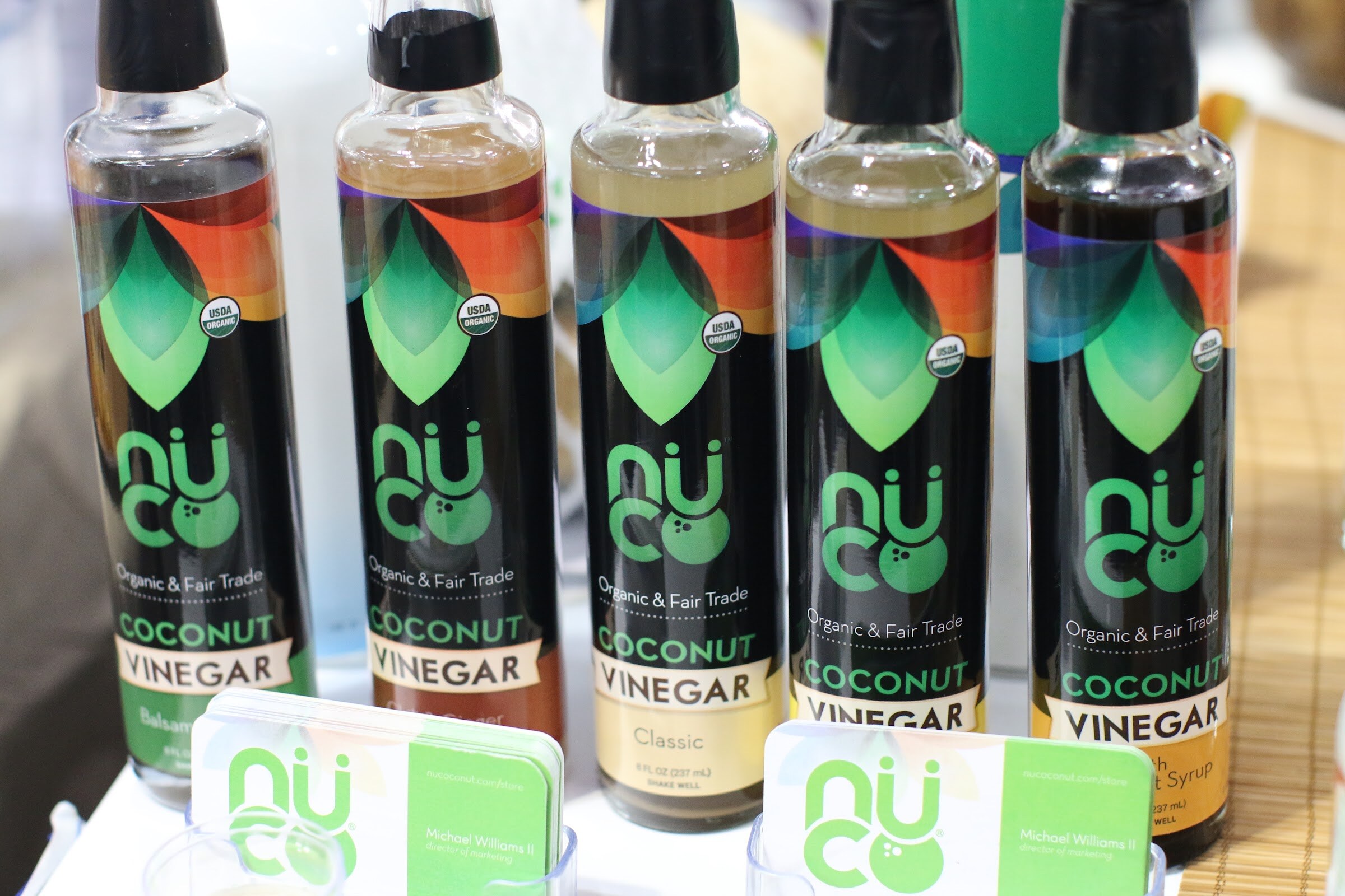 Five tall black NuCo coconut vinegar bottles with green leaf logos above two NuCo business card holders.