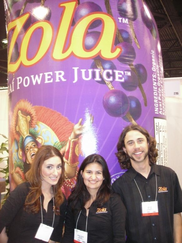 Two women and a man in black Zola shirts and badges stand in front of a giant inflatable Zola Acai Power Juice bottle decorated with acai berries.