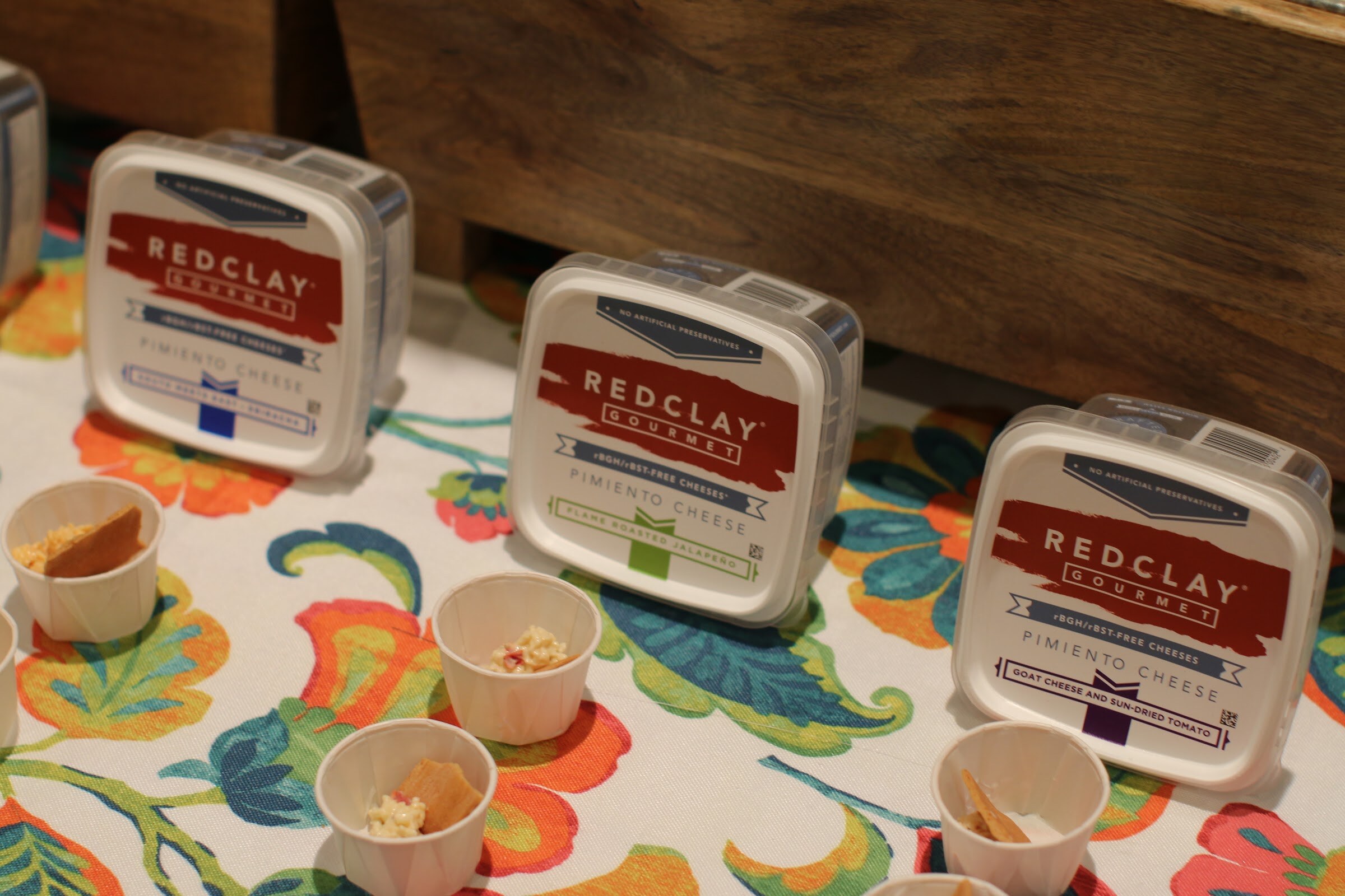 Three tubs of Redclay Gourmet pimiento cheese on a colorful floral tablecloth with paper tasting cups in front.