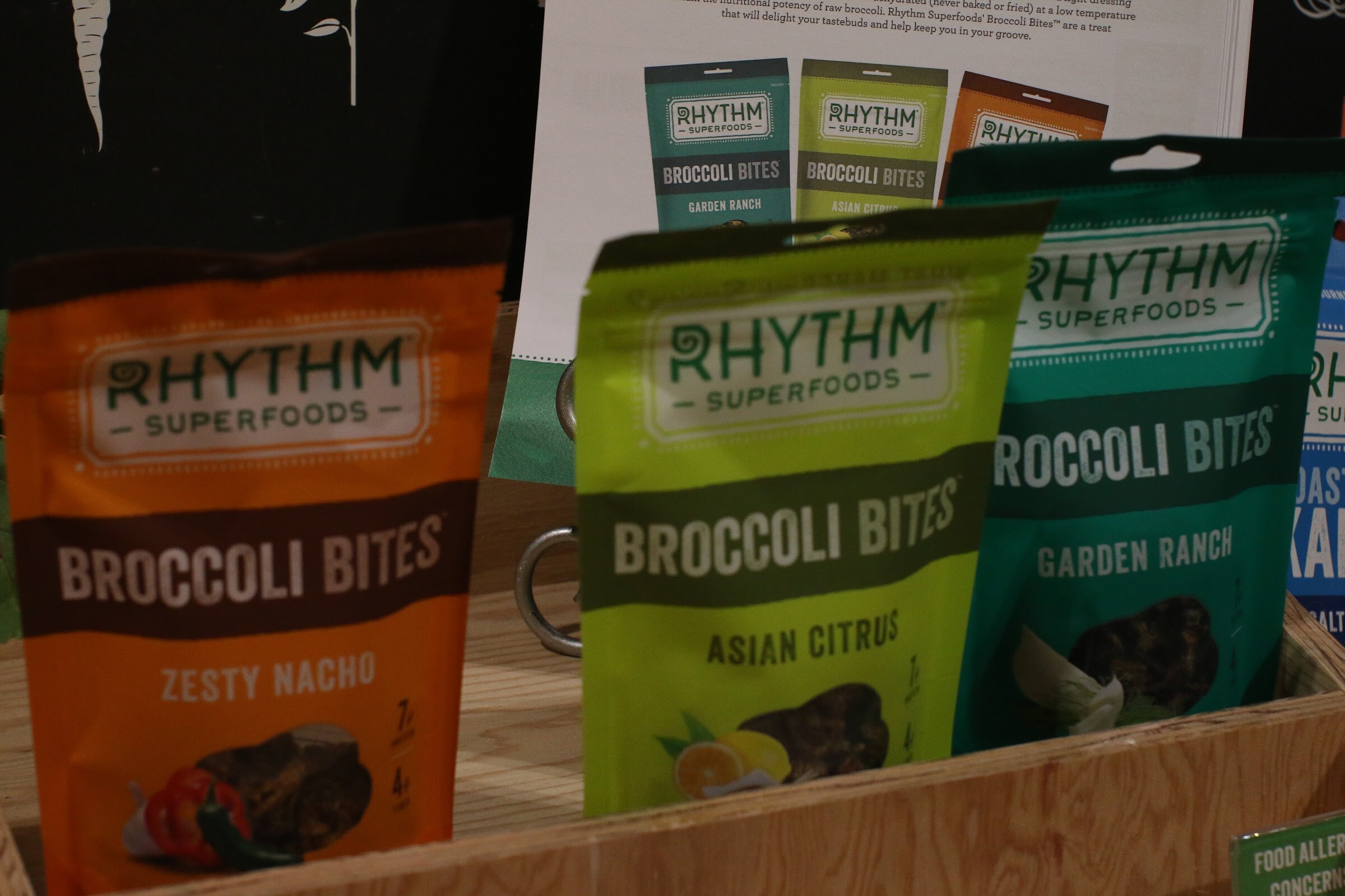 Orange, lime green and teal bags of Rhythm Superfoods broccoli bites in a wooden box.