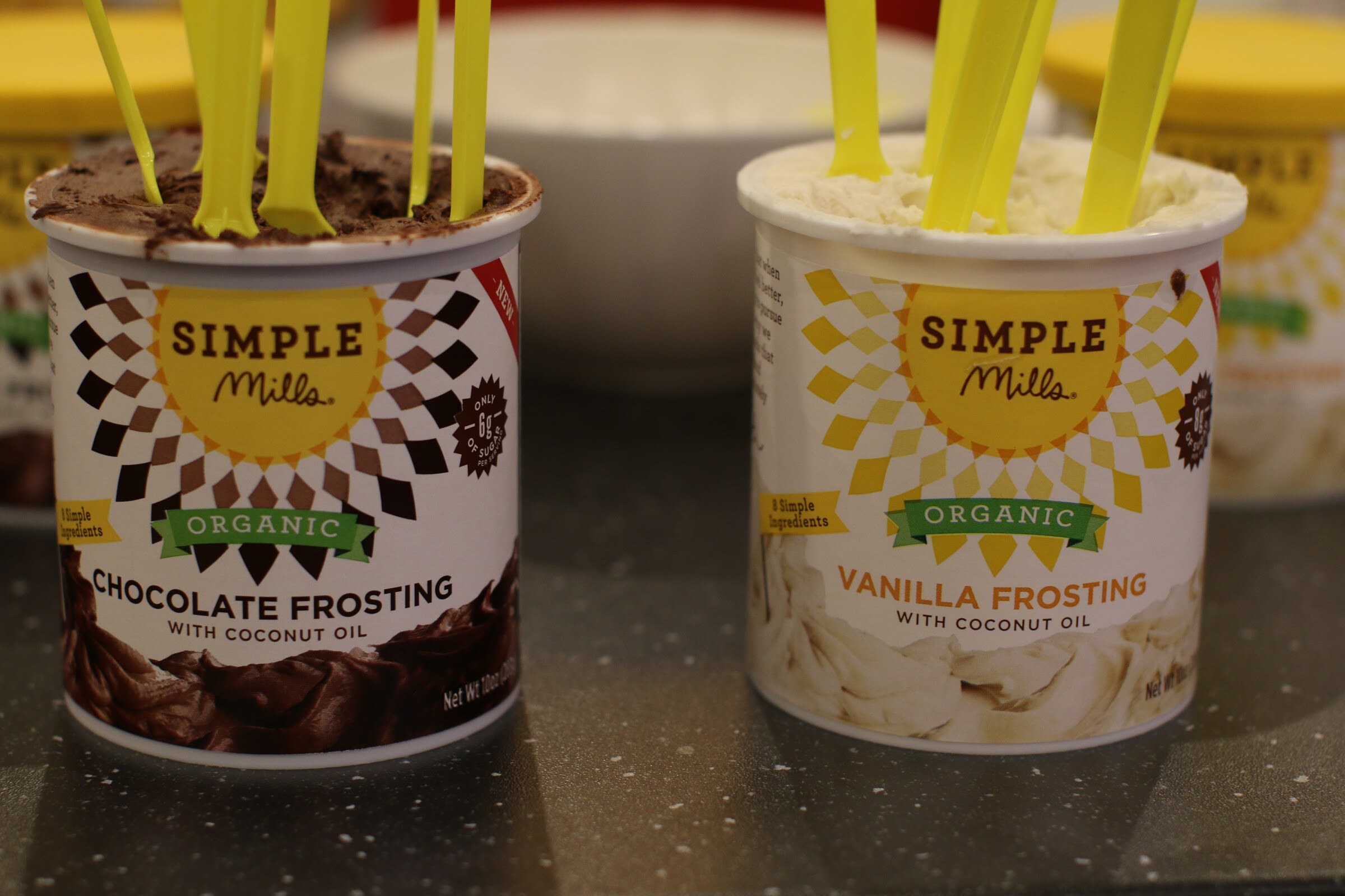Two tubs of Simple Mills organic frosting, chocolate and vanilla, with yellow tasting spoons standing in them.