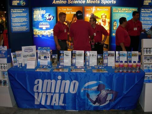 Five people in red shirts stand behind a blue Amino Vital table covered with brochures and bottles, in front of banners reading "Amino Science Meets Sports".