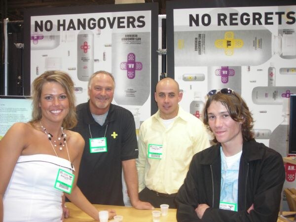 A woman in a white dress and three men stand at a counter with small cups in front of A.H. Anti-Hangover Beverage posters reading No Hangovers and No Regrets.