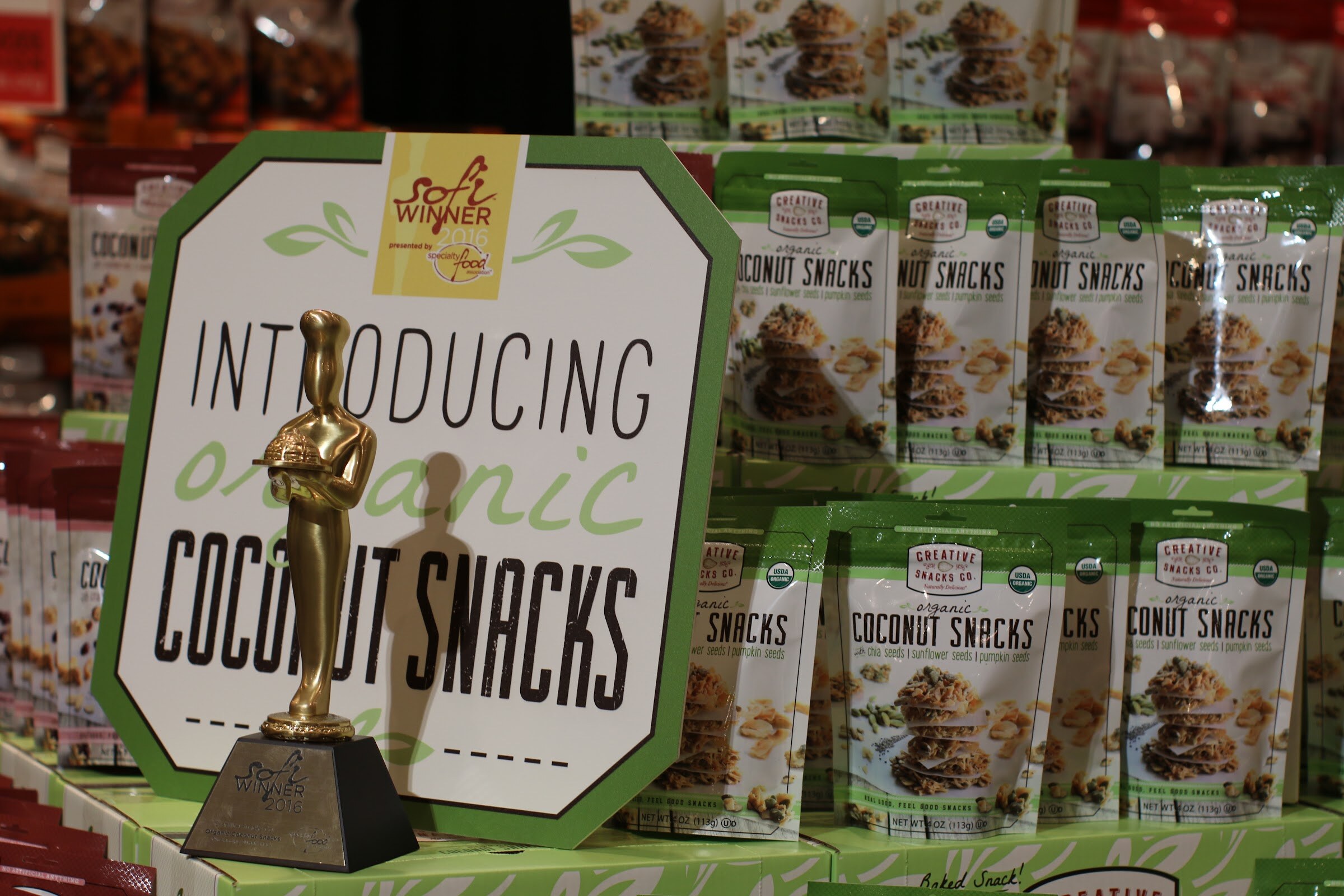 Gold chef-shaped award on a black base next to a green and white sign for organic coconut snacks, with rows of snack bags behind.