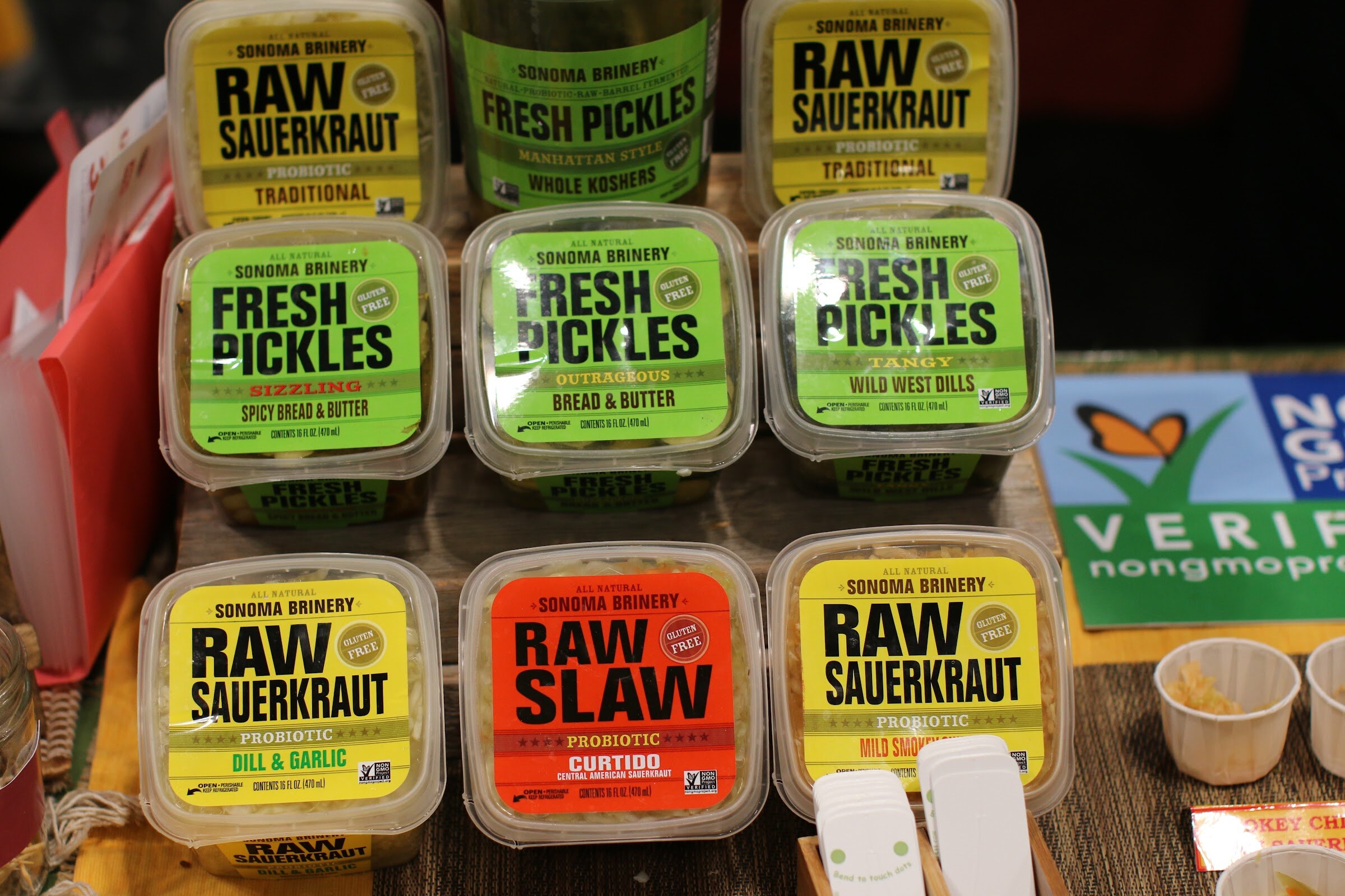 Tubs of Sonoma Brinery raw sauerkraut and fresh pickles in yellow, green and red labels with a Non-GMO Project sign beside them.