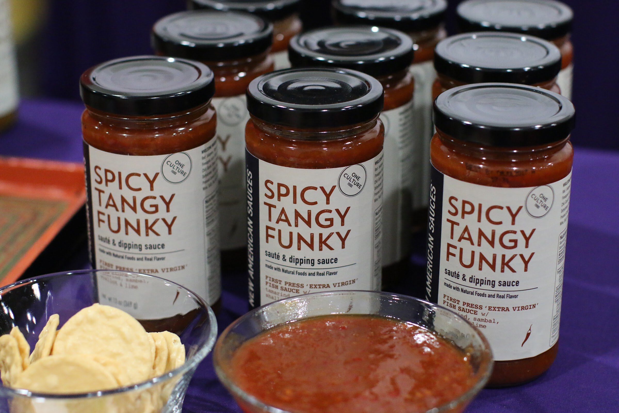 Several jars of Spicy Tangy Funky sauce on a purple tablecloth with a bowl of sauce and tortilla chips.