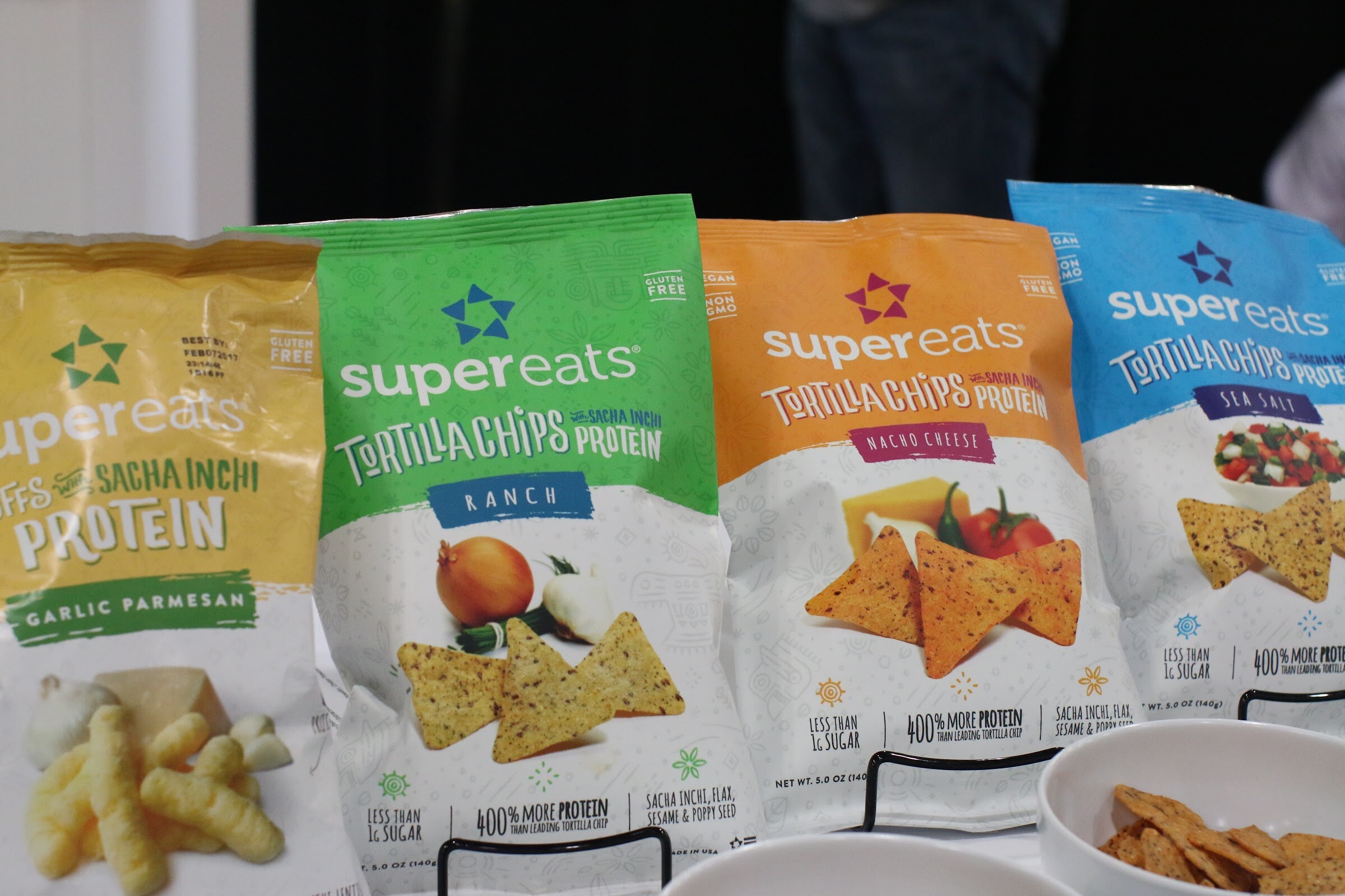 Four Supereats snack bags in yellow, green, orange and blue standing on black wire stands next to a bowl of chips.