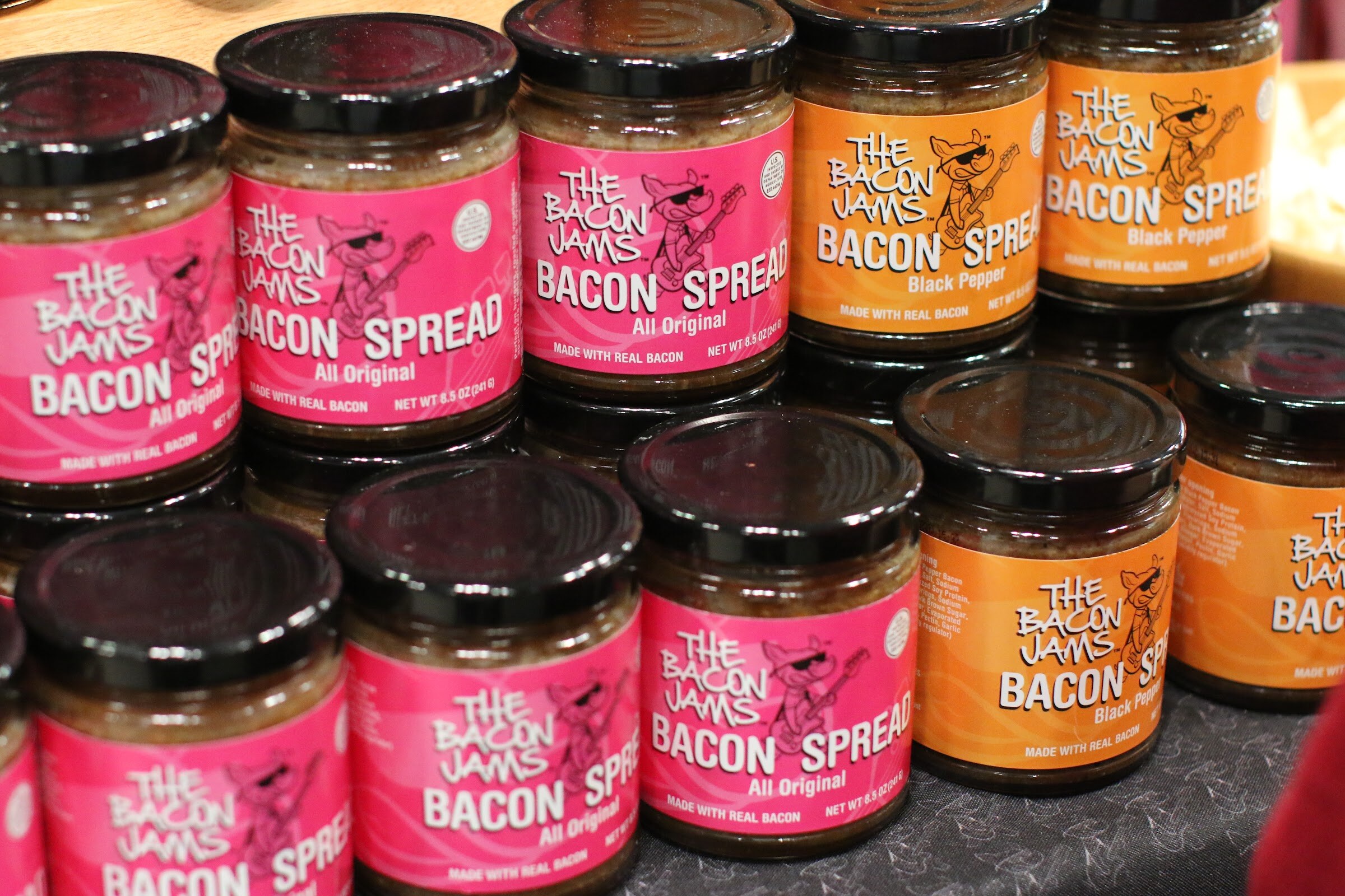 Stacked jars of The Bacon Jams bacon spread with pink and orange labels and black lids.