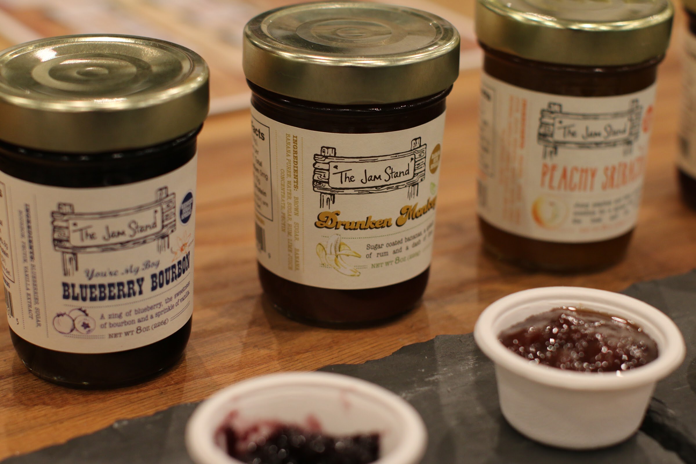 Three jars of The Jam Stand jam on a wood table with two small white cups of jam on a slate board.