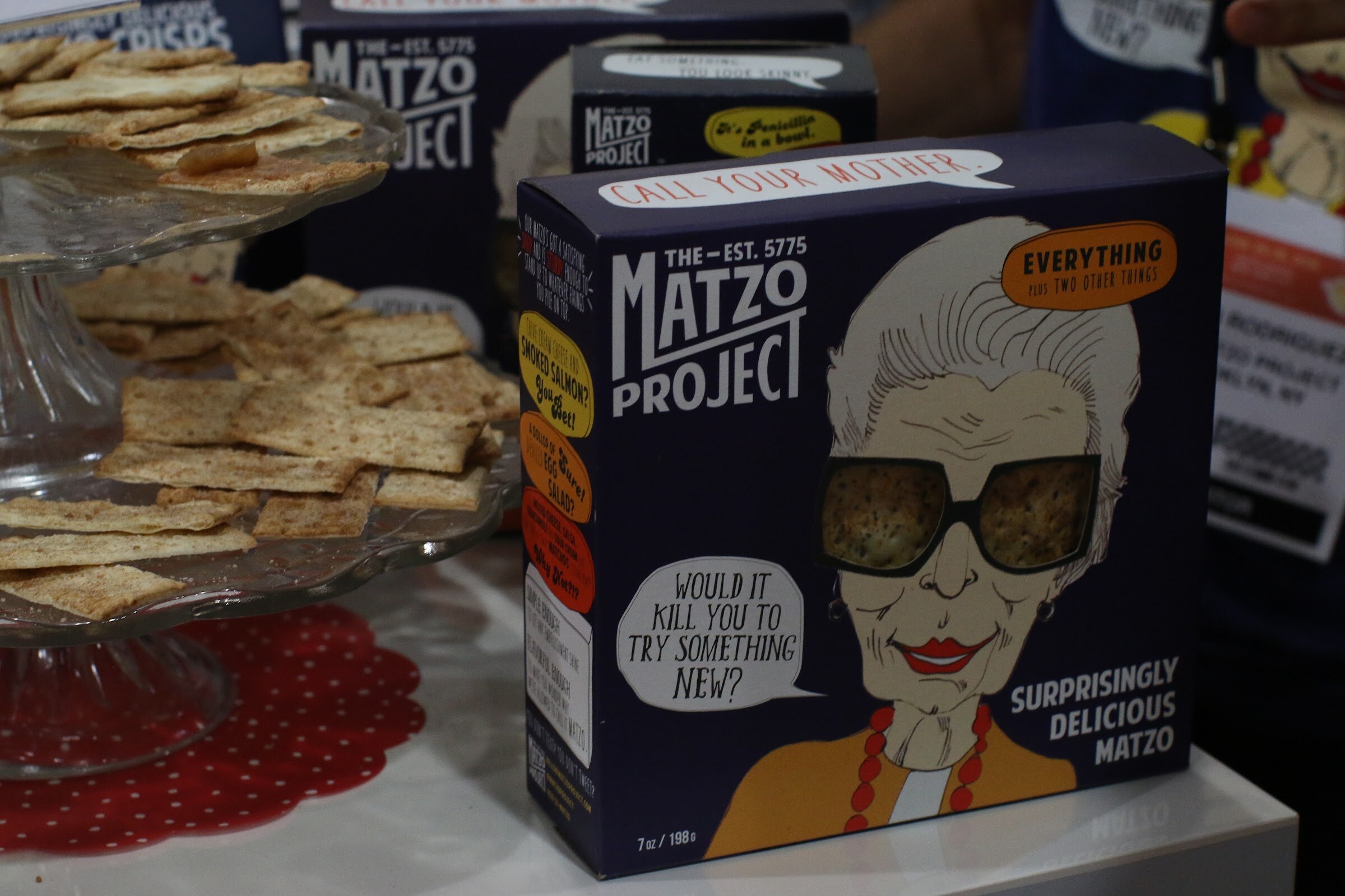 Navy The Matzo Project box with a cartoon of an older woman in sunglasses, beside a glass stand of matzo crackers.