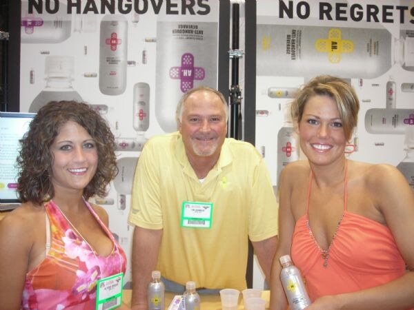 A man in a yellow polo stands between two smiling women, one holding a bottle, at a counter with cups and bottles before A.H. Anti-Hangover Beverage posters.