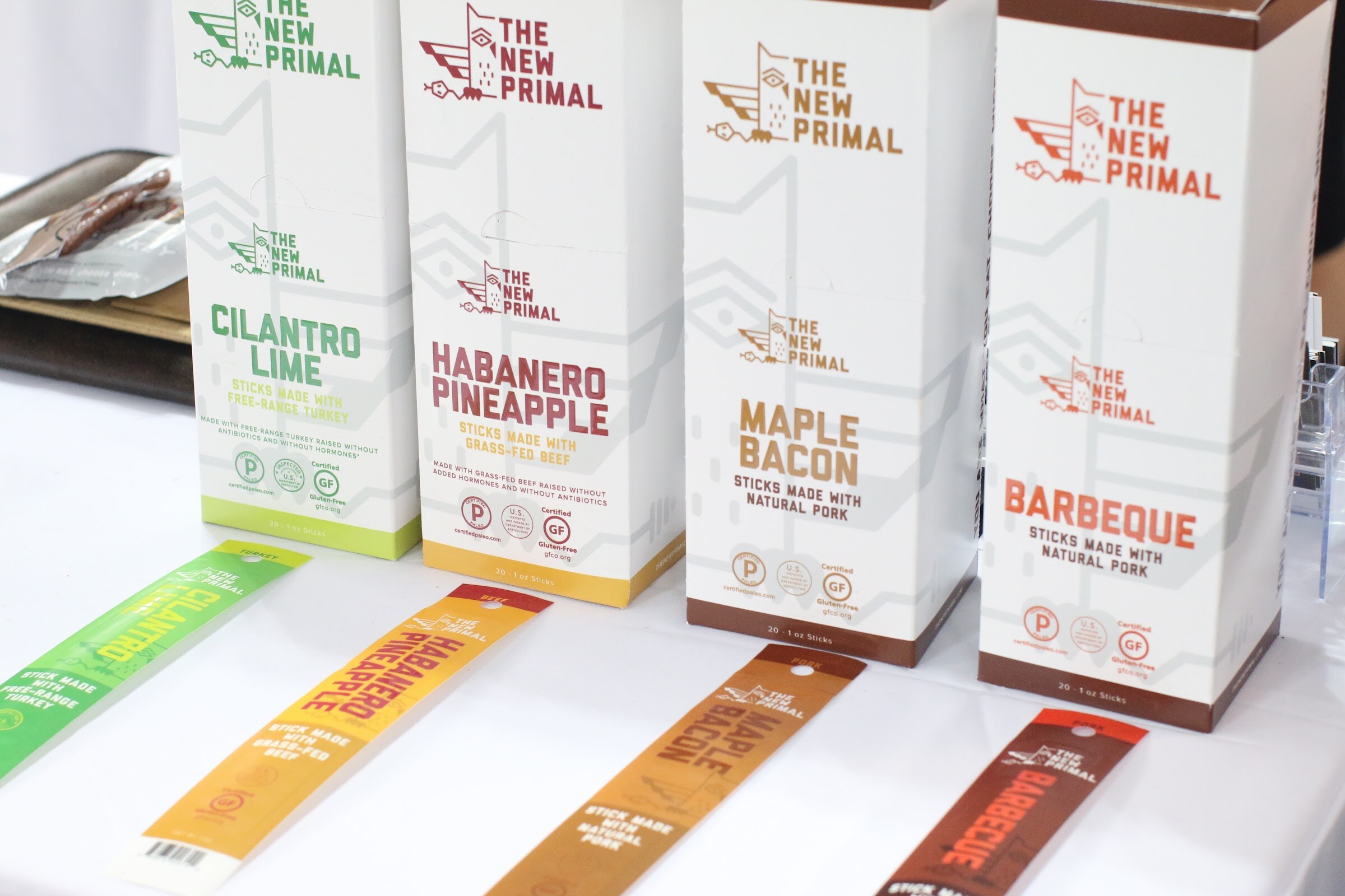 Four white The New Primal boxes with colored flavor names and four single meat stick wrappers laid out below them.