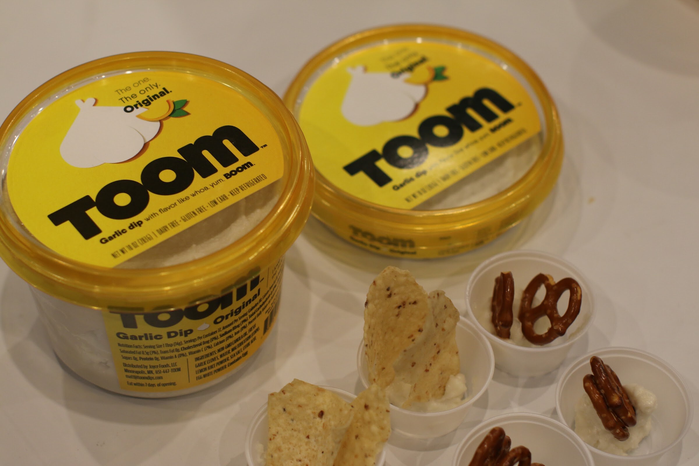 Two yellow tubs of TOOM garlic dip beside small cups of dip with pretzels and tortilla chips.