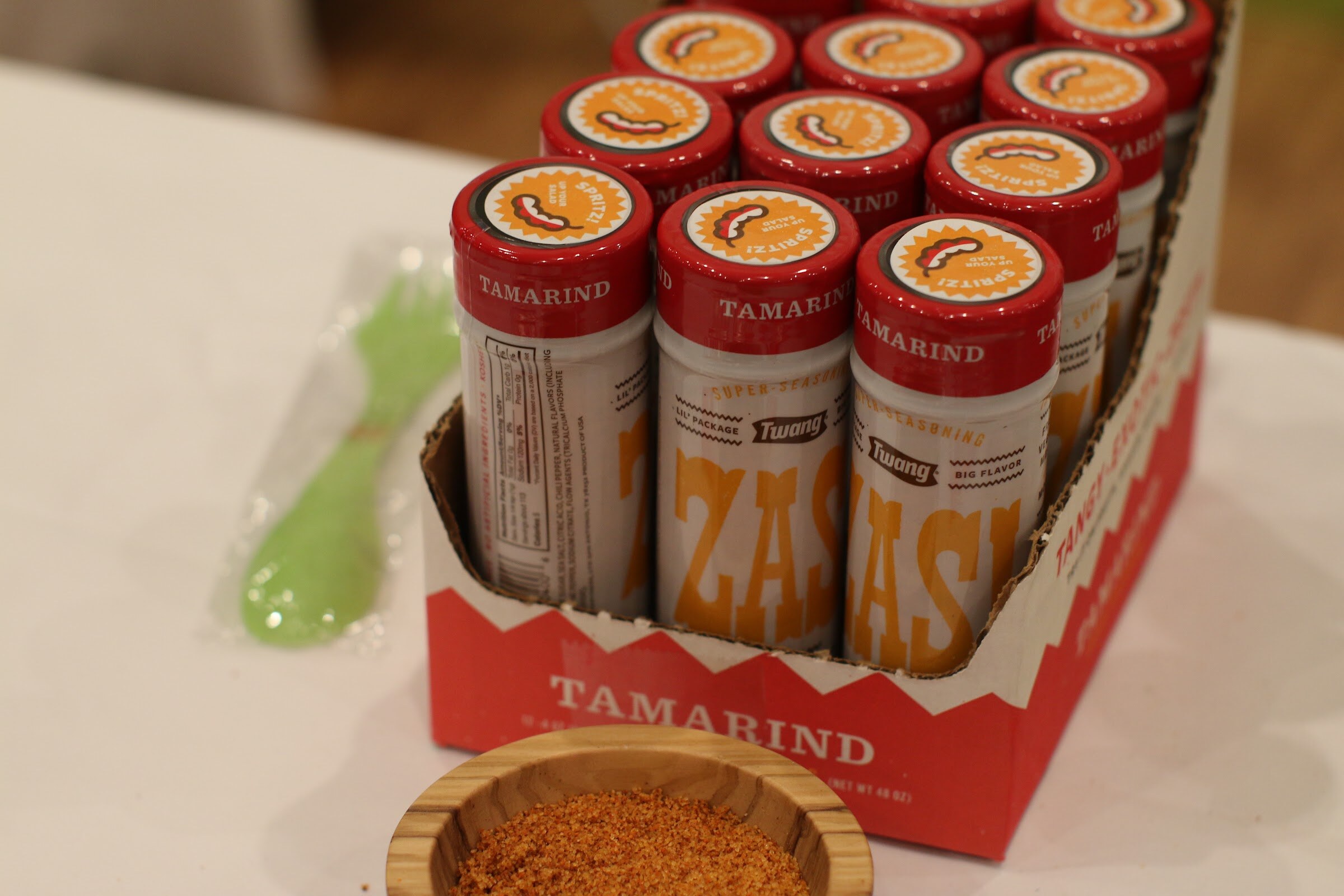 Open red cardboard box of tamarind seasoning bottles with a bowl of orange-red seasoning and a green utensil beside it.